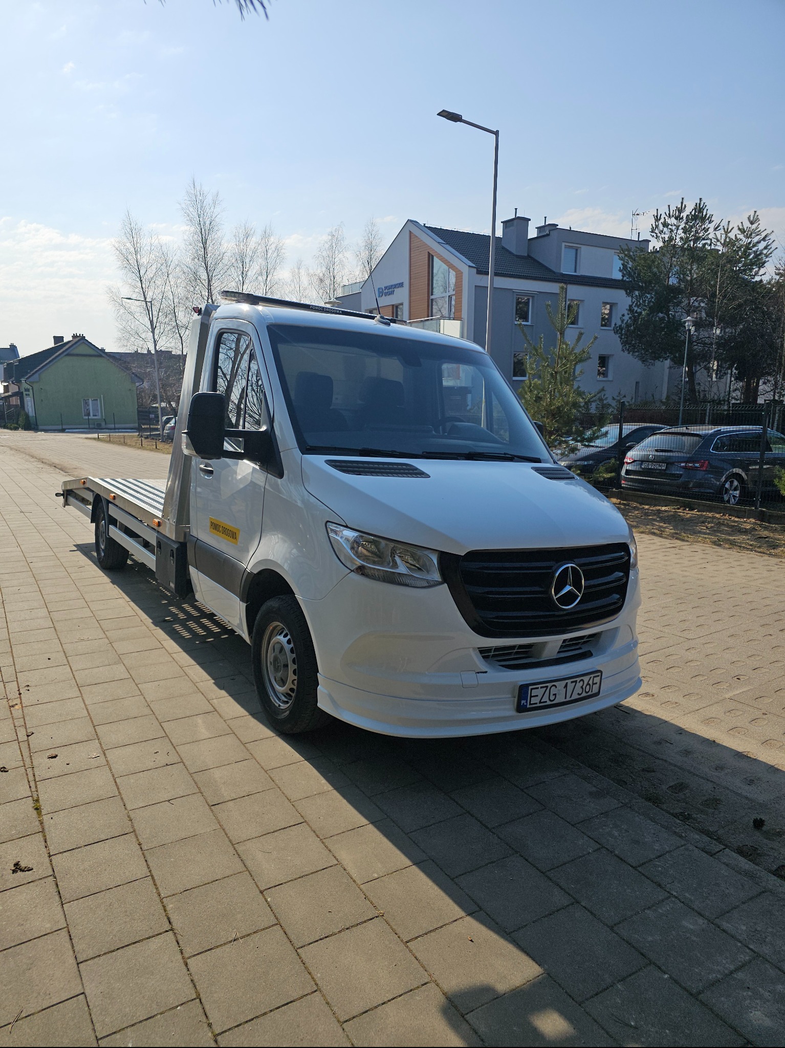 Biały Mercedes-Benz Sprinter z platformą, zaparkowany na chodniku w słoneczny dzień. Widoczny napis 'Pomoc Drogowa' na boku pojazdu i budynki w tle.