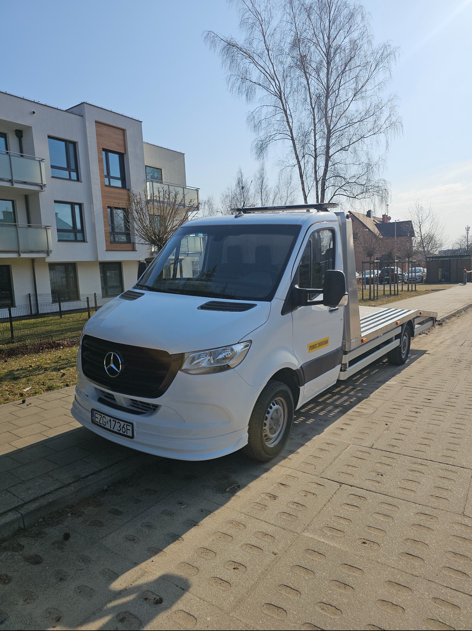 Biała autolaweta Mercedes na chodniku, z napisem 'Pomoc Drogowa' na boku, zaparkowana przed nowoczesnym budynkiem mieszkalnym w słoneczny dzień.
