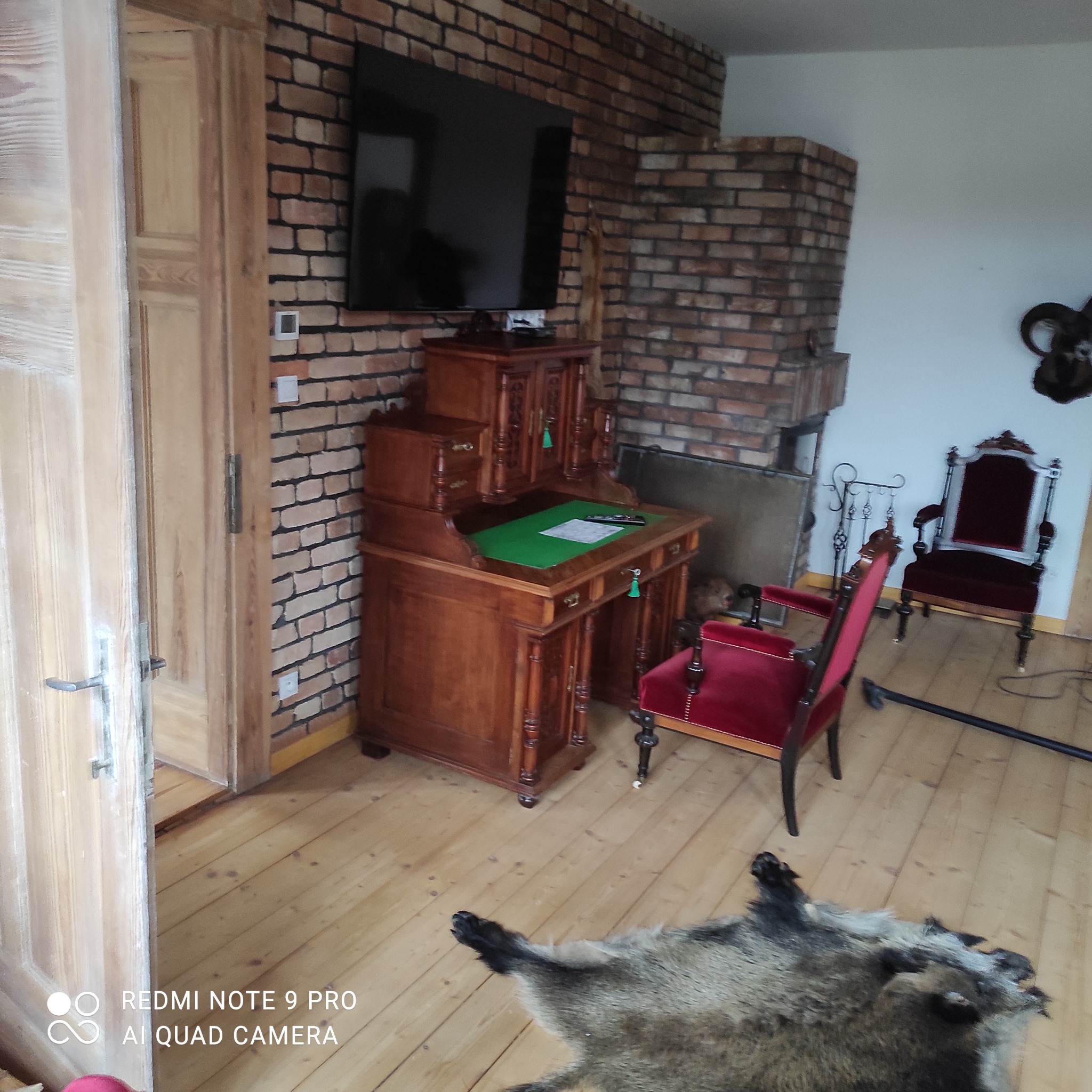 Antyczny gabinet z zielonym blatem przy ceglanej ścianie z telewizorem, obok czerwone krzesła i skóra dzika na drewnianej podłodze.