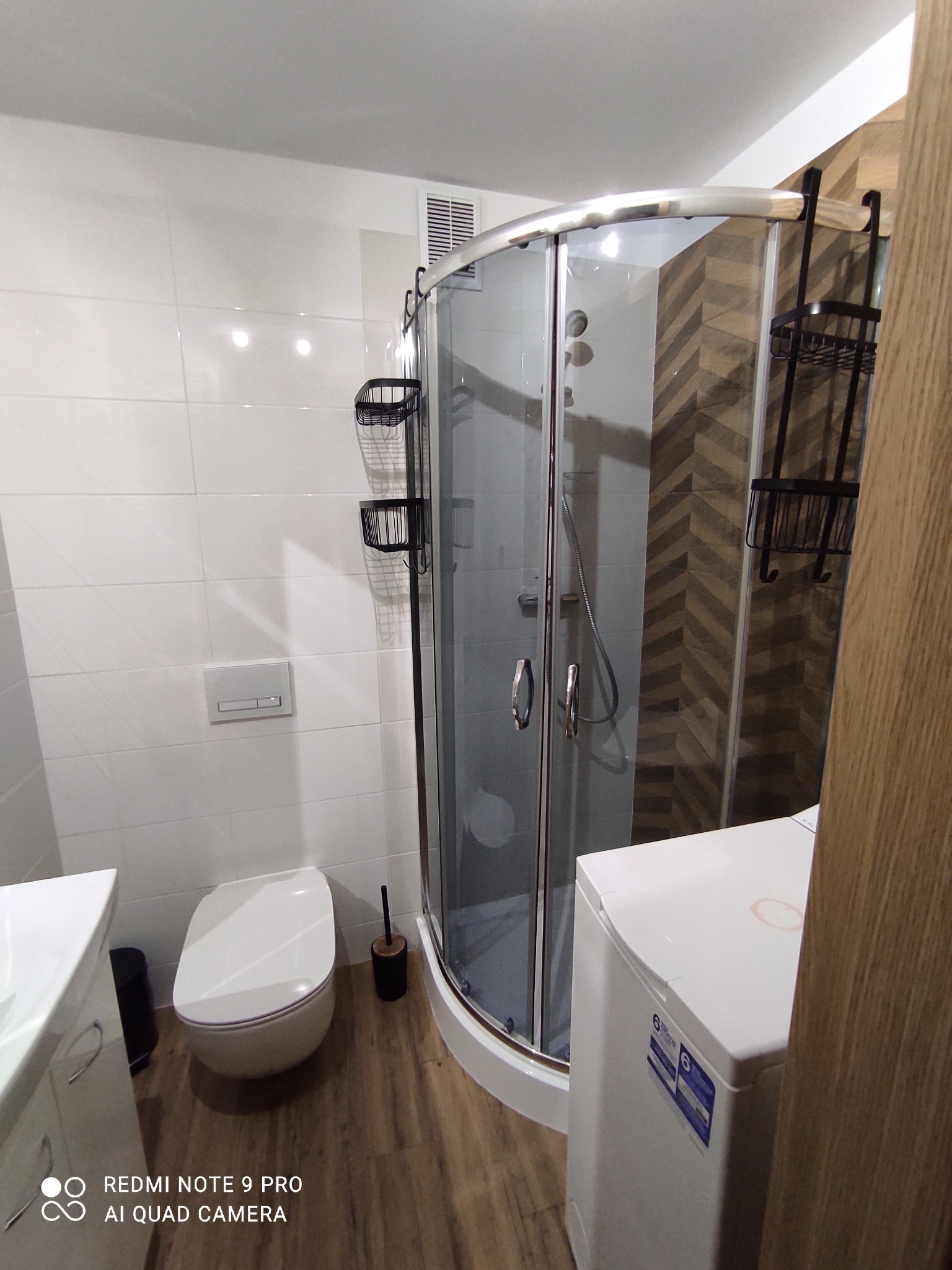 Nowoczesna łazienka z kabiną prysznicową, białą toaletą, umywalką i pralką z widoczną plamą na górnej obudowie.