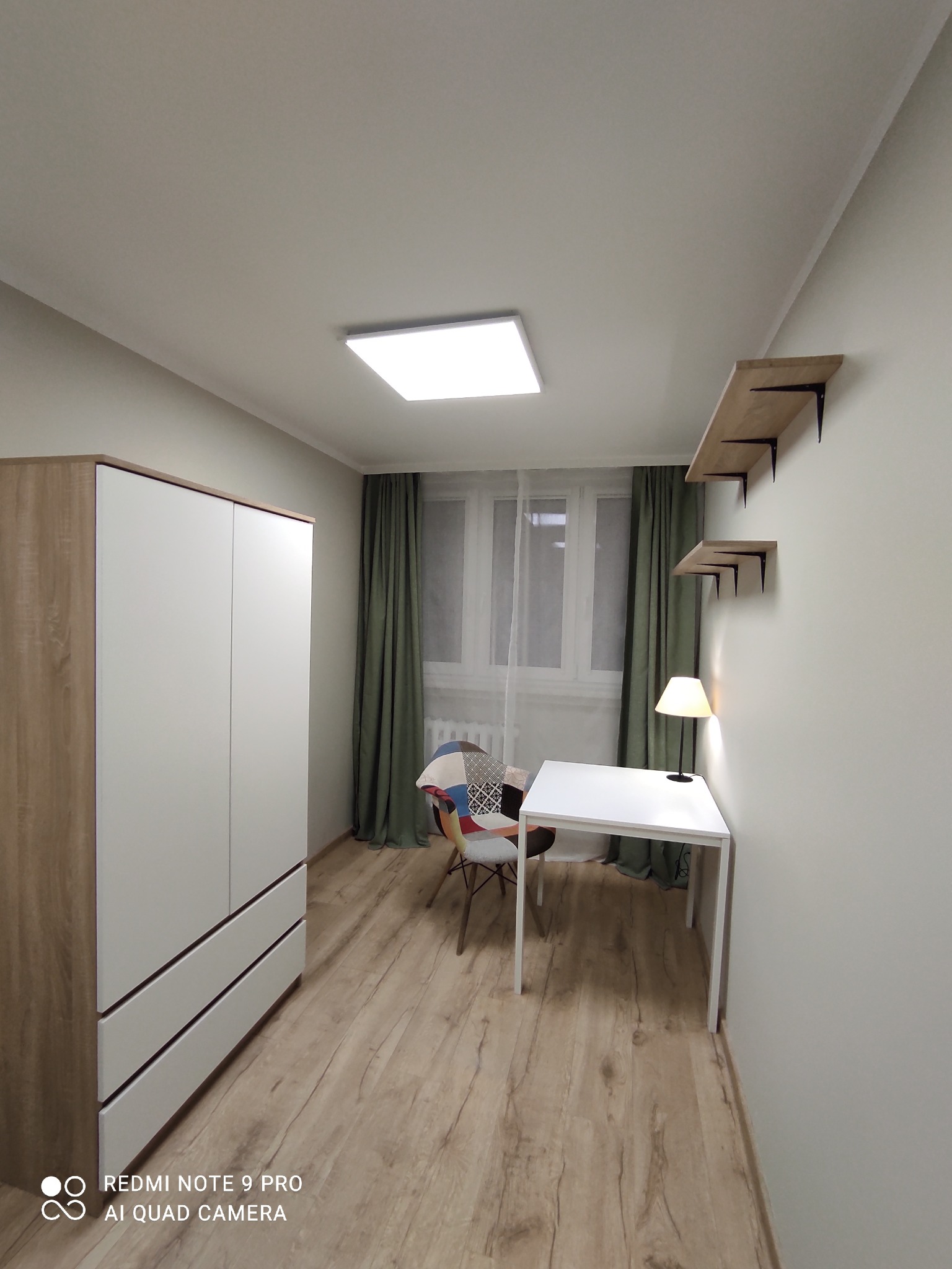 Pokój po odświeżeniu: biała szafa z drewnianymi bokami, biurko z lampką, krzesło patchworkowe, półki na ścianie, drewniana podłoga i zielone zasłony w oknie.