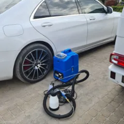 Niebieski odkurzacz piorący obok białych samochodów Mercedes klasy S i G, przygotowany do czyszczenia tapicerki samochodowej.