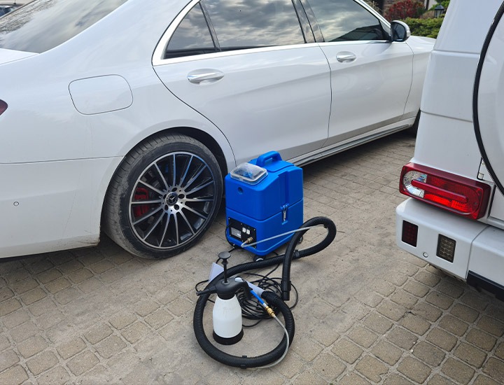 Niebieski odkurzacz piorący obok białych samochodów Mercedes klasy S i G, przygotowany do czyszczenia tapicerki samochodowej.