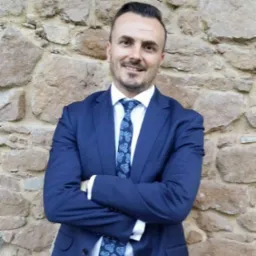 Mężczyzna w niebieskim garniturze i wzorzystym krawacie, z założonymi rękami, pozuje na tle ściany z naturalnego kamienia.