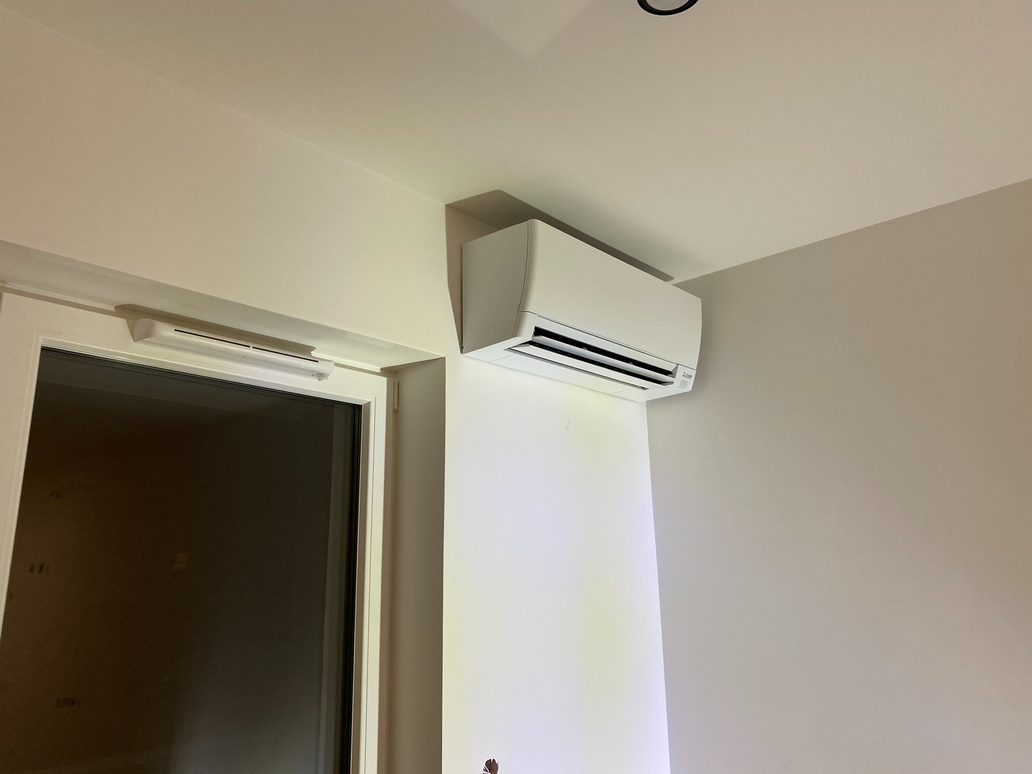 Biały klimatyzator ścienny zamontowany w pomieszczeniu w Rumi, widoczny obok okna z ciemną szybą, na tle jasnych ścian.