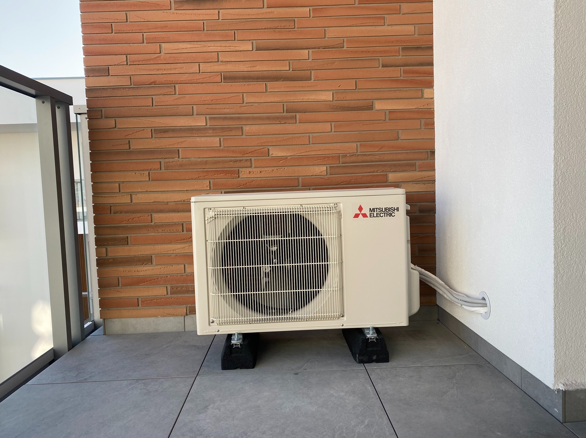 Jednostka zewnętrzna klimatyzacji Mitsubishi Electric zamontowana na balkonie z ceglaną ścianą w tle, widoczne kable wchodzące do otworu w ścianie.