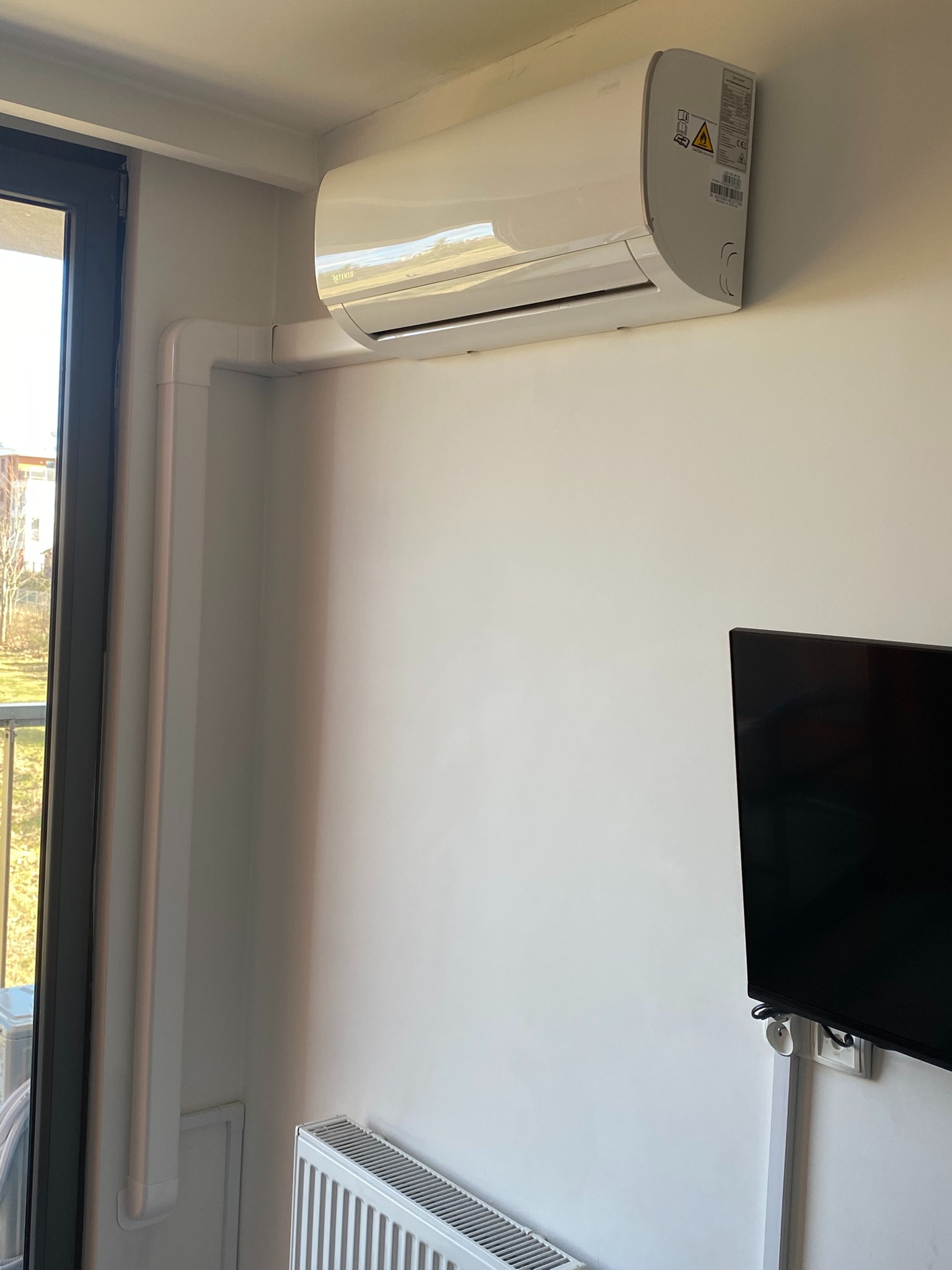 Biały klimatyzator ścienny zamontowany w jasnym pomieszczeniu, z widocznymi rurami odprowadzającymi i fragmentem telewizora na ścianie.