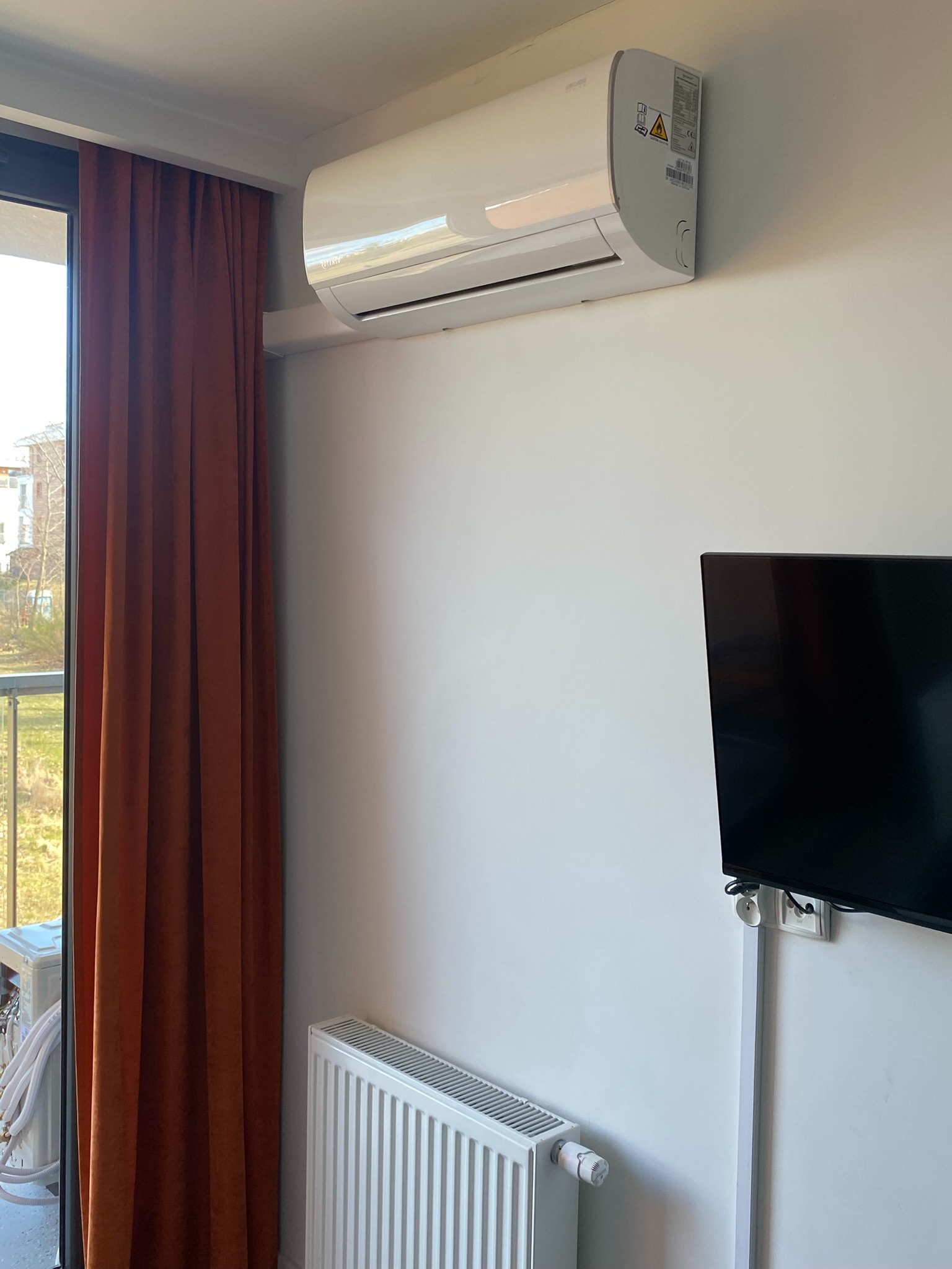 Biały klimatyzator zamontowany na ścianie w pokoju z telewizorem i grzejnikiem, pomarańczowe zasłony w tle.