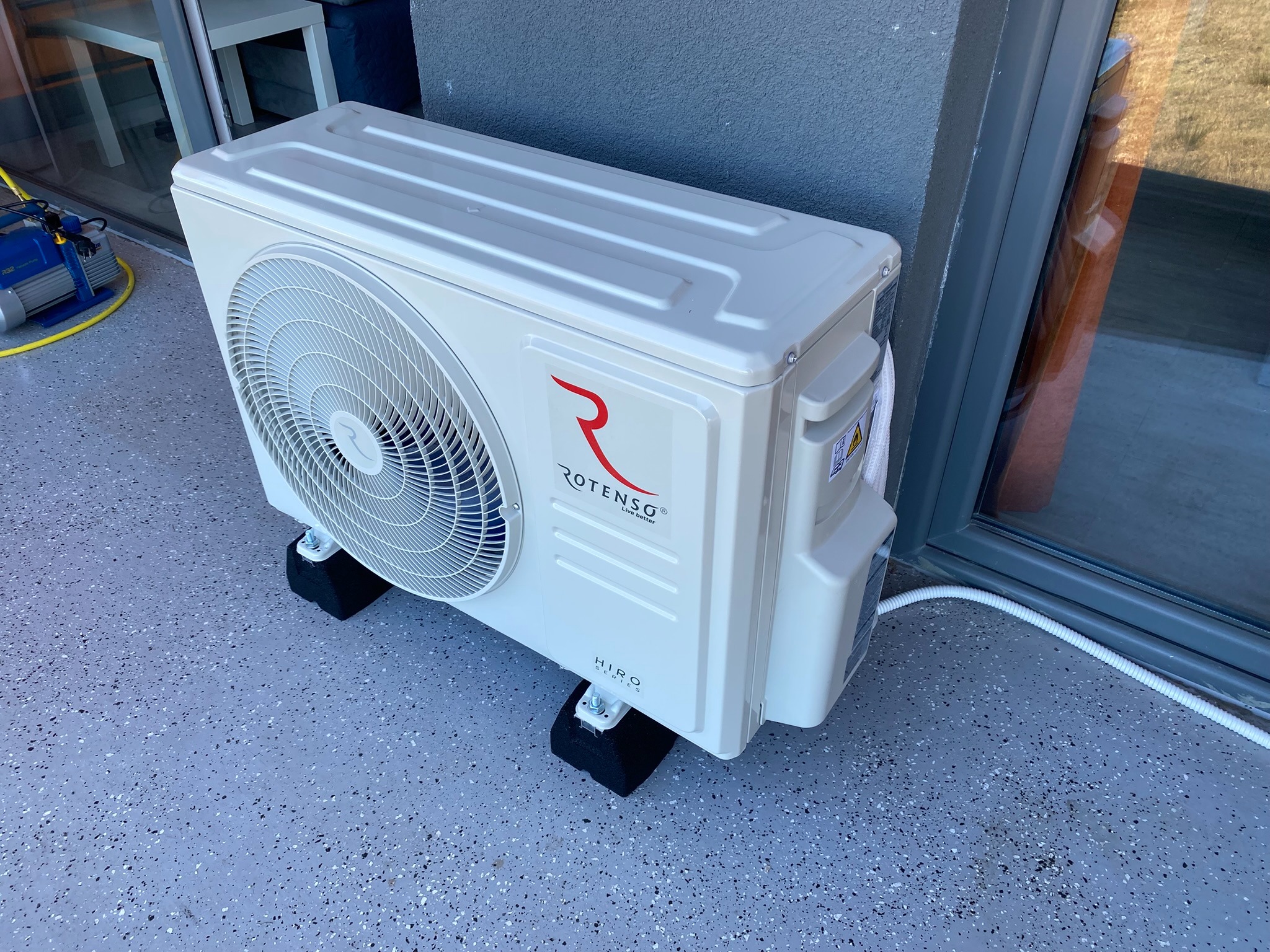 Zewnętrzna jednostka klimatyzacji Rotenso Hiro Series, zamontowana na czarnych podkładkach antywibracyjnych na szarej posadzce z żywicy epoksydowej, z widoczną białą rurą odprowadzającą skropliny...