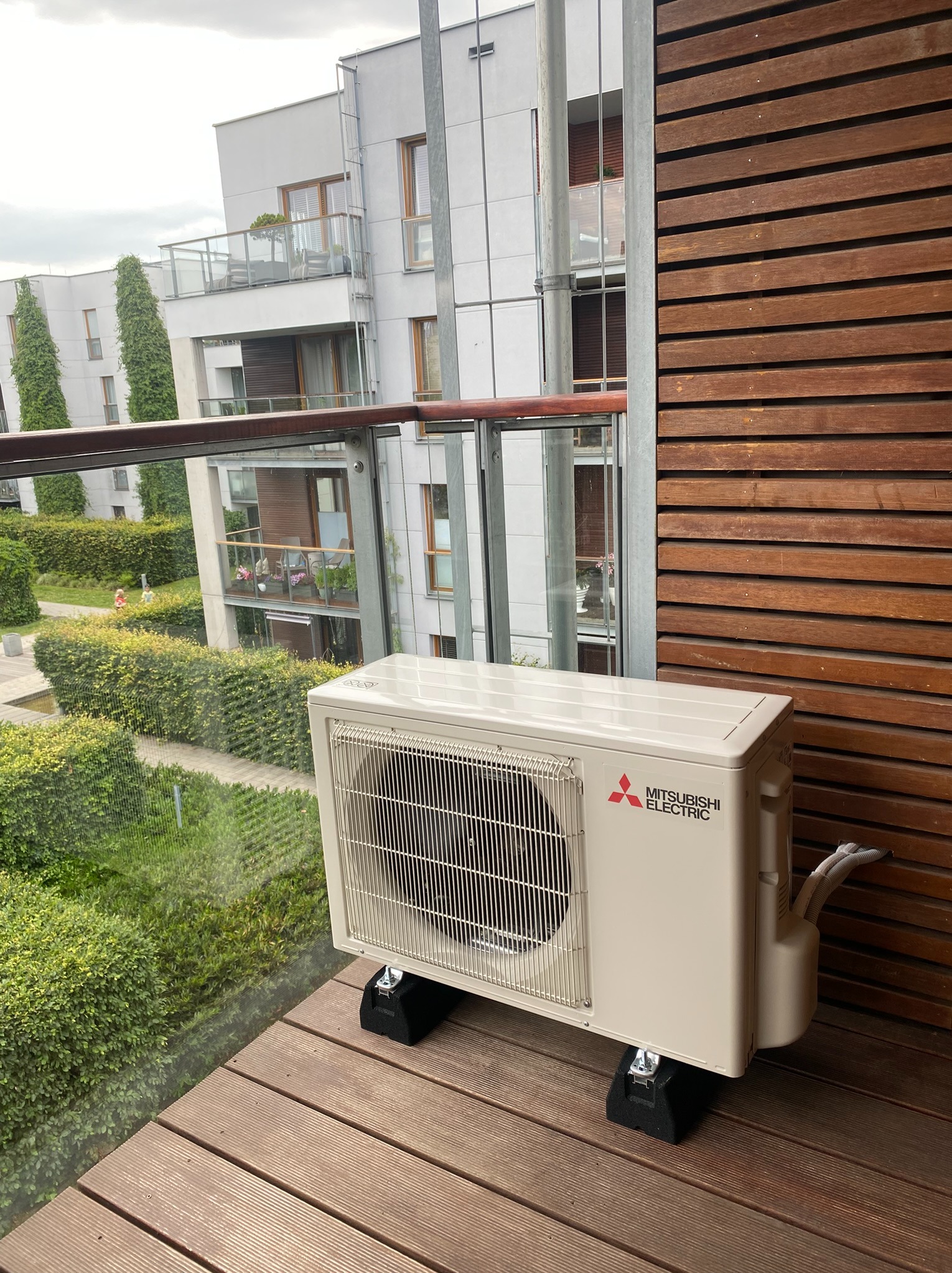 Klimatyzator Mitsubishi Electric na balkonie z drewnianą podłogą i ścianą. Widok na budynek mieszkalny i zieleń. Montaż na specjalnych podkładkach antywibracyjnych.