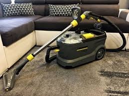 Ekstraktor do czyszczenia dywanów Karcher Puzzi z akcesoriami na szarym dywanie, w tle narożnik z ciemną tapicerką i wzorzystymi poduszkami.