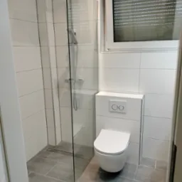 Nowoczesna łazienka z kabiną prysznicową typu walk-in, podwieszaną toaletą i szarymi płytkami podłogowymi, widok na okno z roletami.