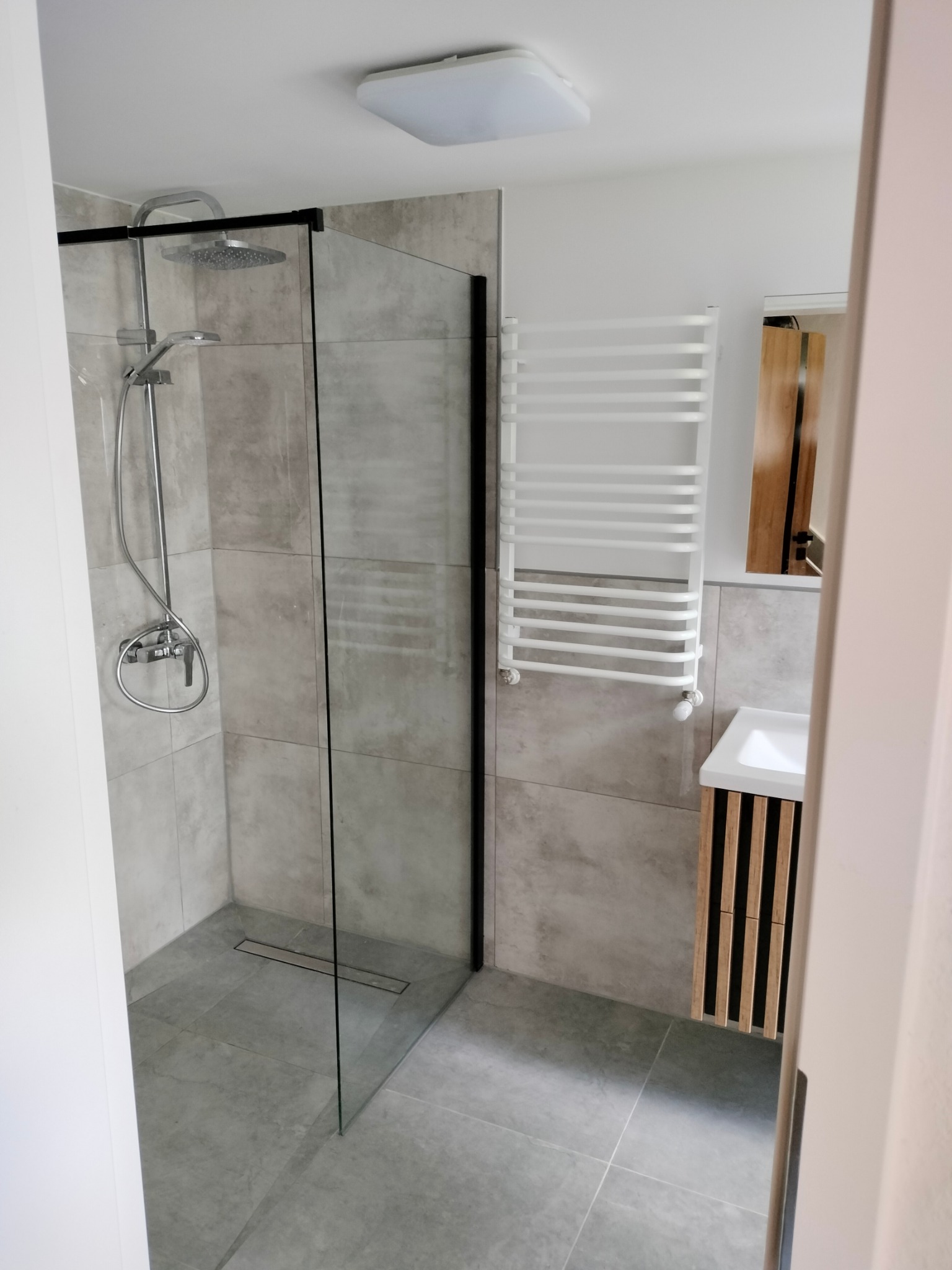 Nowoczesna łazienka z kabiną prysznicową typu walk-in z czarnymi profilami, grzejnikiem drabinkowym i umywalką z szafką z lamelami.