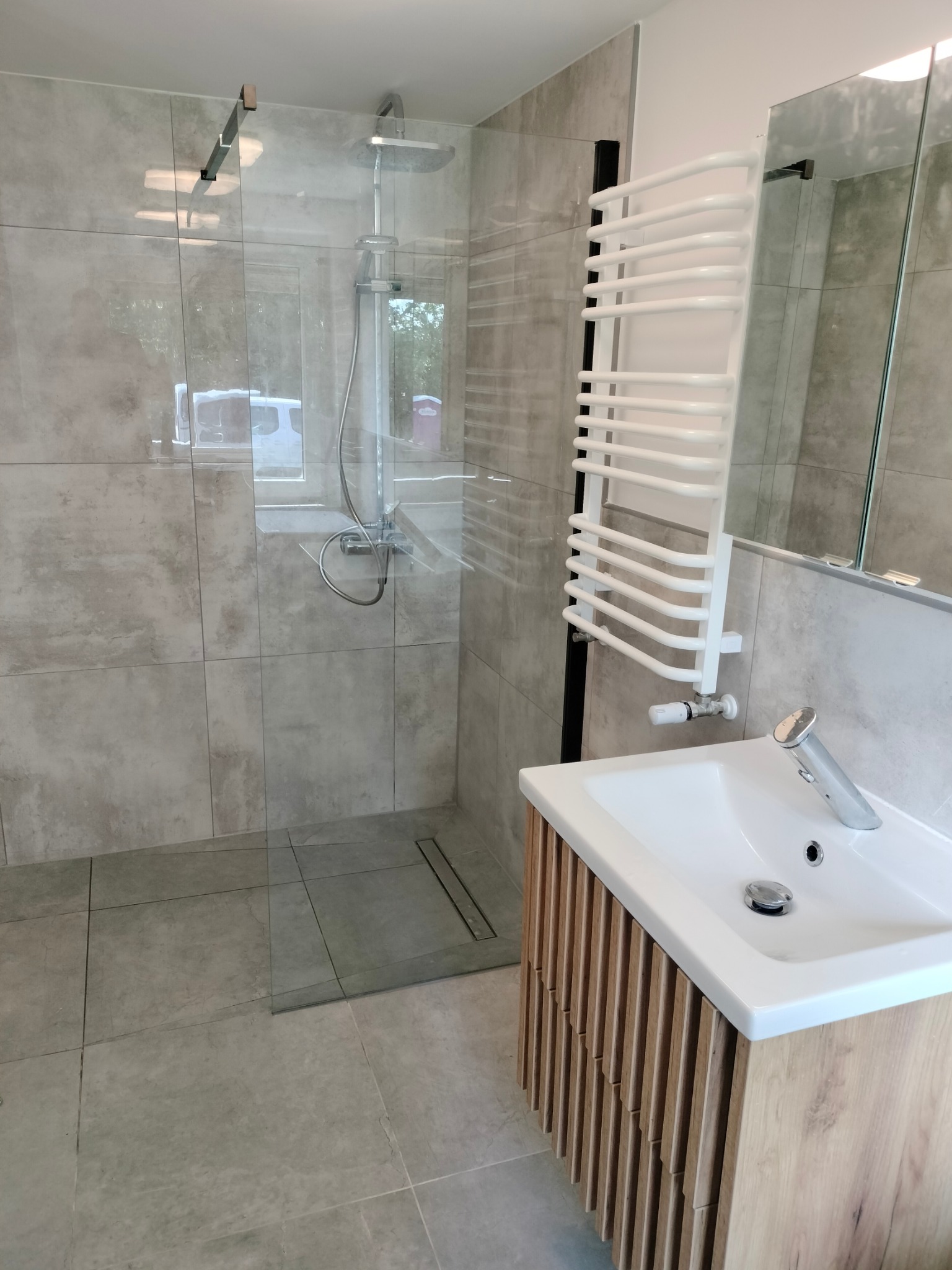 Nowoczesna łazienka z kabiną prysznicową typu walk-in, grzejnikiem drabinkowym, umywalką nablatową na szafce z lamelami i dużym lustrem.