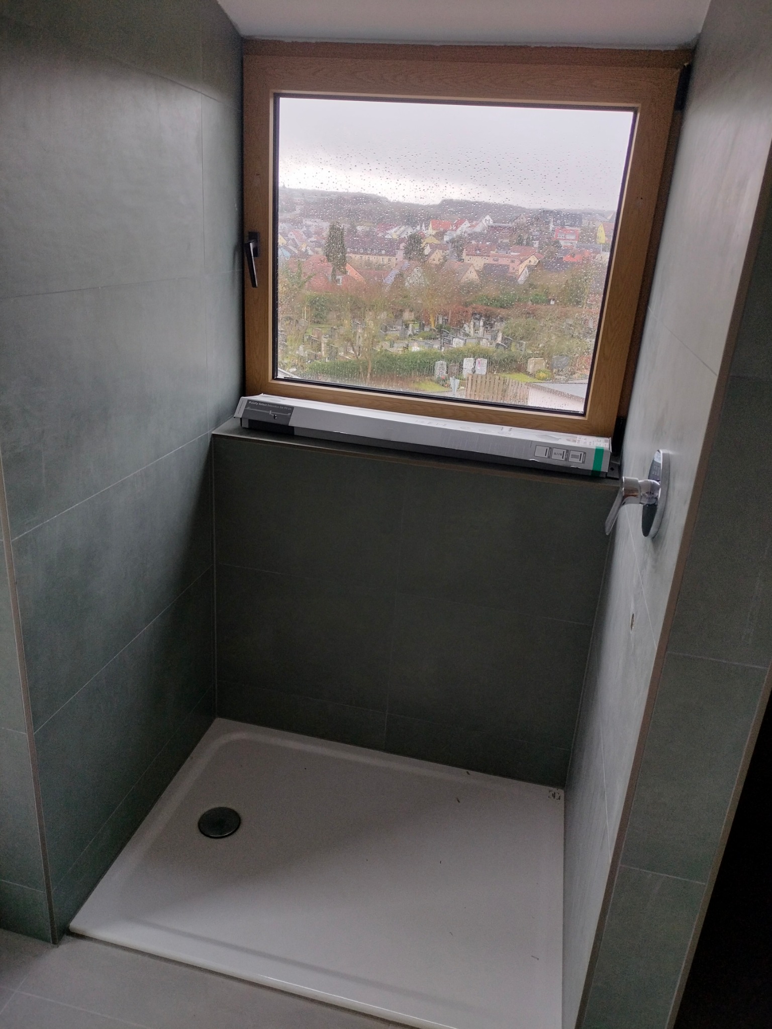 Szara kabina prysznicowa z oknem dachowym z widokiem na miasto, bateria zamontowana na ścianie, opakowanie po baterii leży na parapecie.