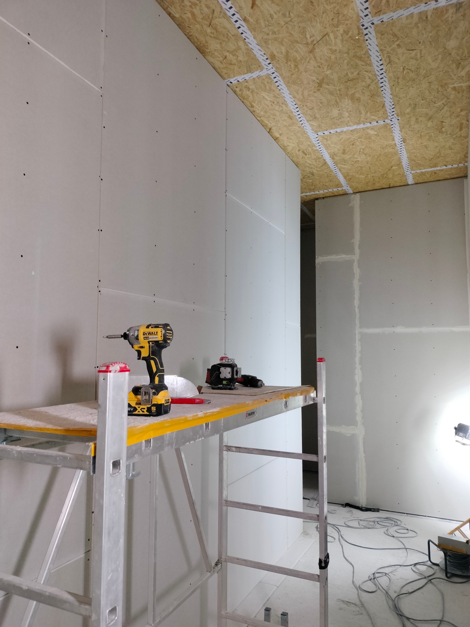 Montaż płyt gipsowo-kartonowych na ścianie, widoczny wkrętak DeWalt na rusztowaniu, laser krzyżowy oraz sufit z płyt OSB z metalowymi profilami.
