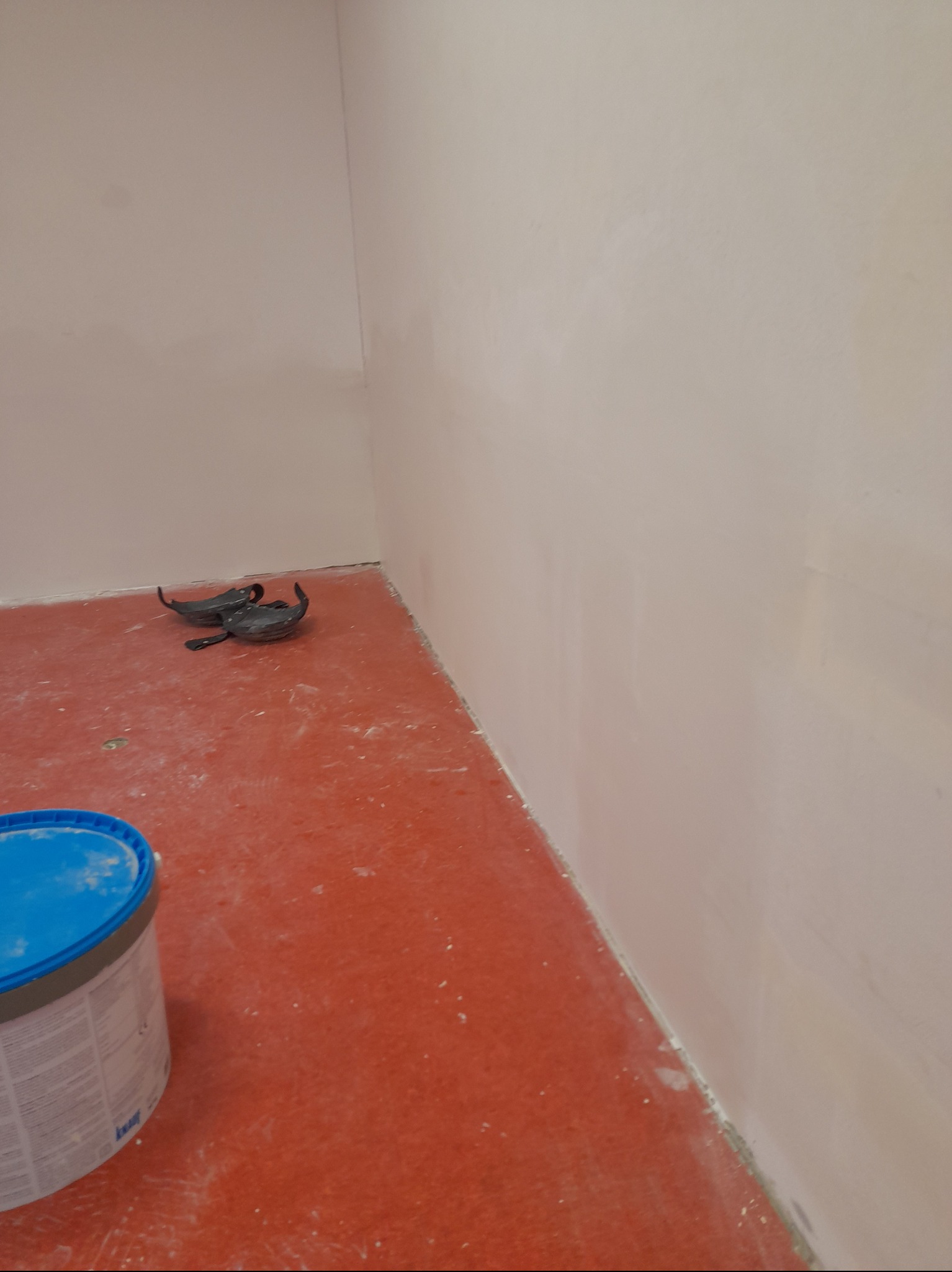 Ściana po szpachlowaniu, widoczny kubeł z masą szpachlową i narzędzia na czerwonej podłodze. Świeżo wykończona powierzchnia gotowa do malowania.