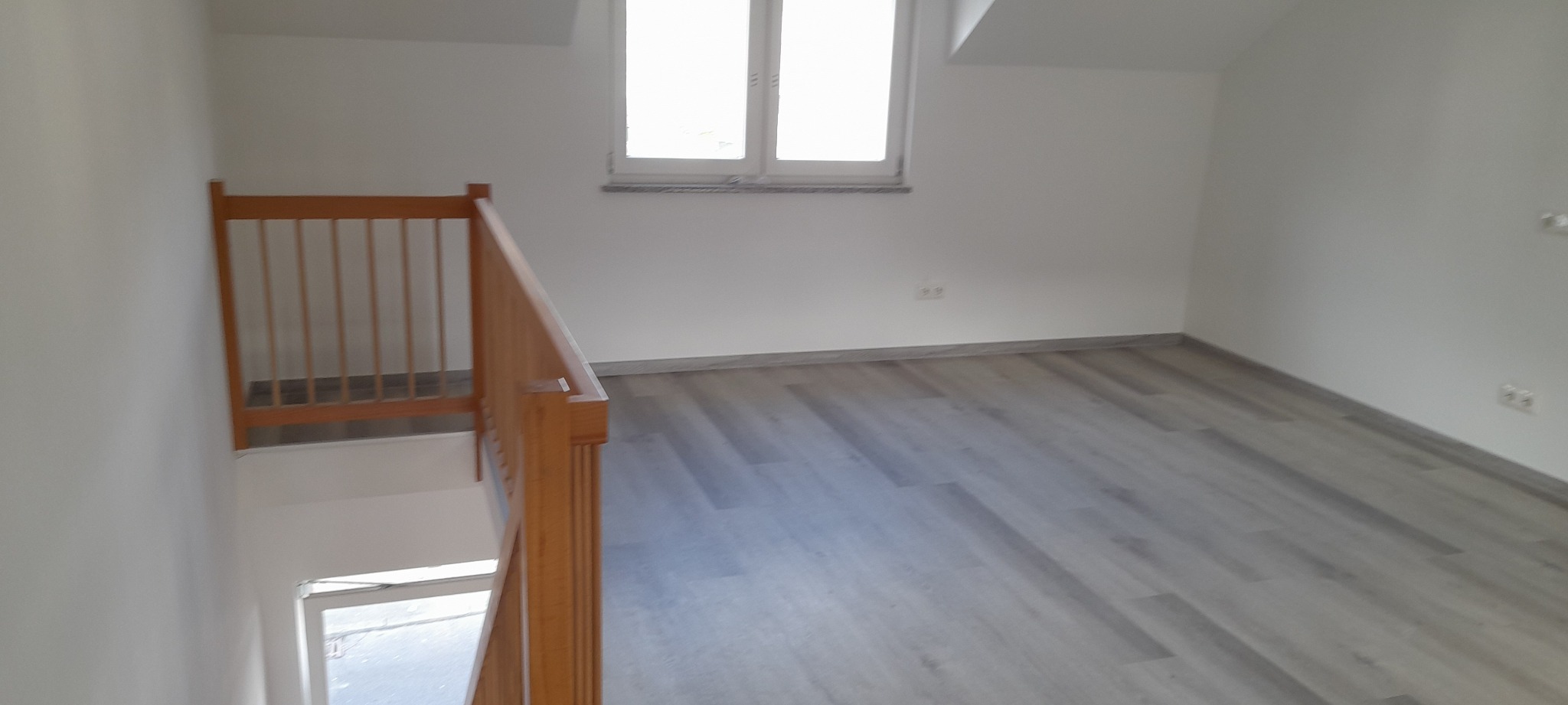 Jasne panele podłogowe w nowym pokoju na poddaszu, widok z góry schodów z drewnianą barierką. Świeżo pomalowane ściany i okno dachowe.