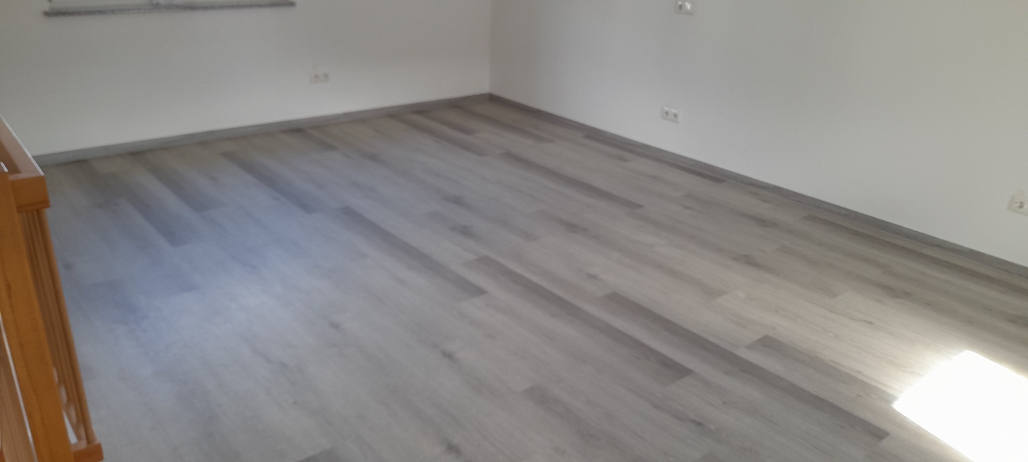 Jasnoszara podłoga z paneli ułożona w pustym, odnowionym pokoju z białymi ścianami i gniazdkami elektrycznymi. Fragment drewnianej barierki po lewej.