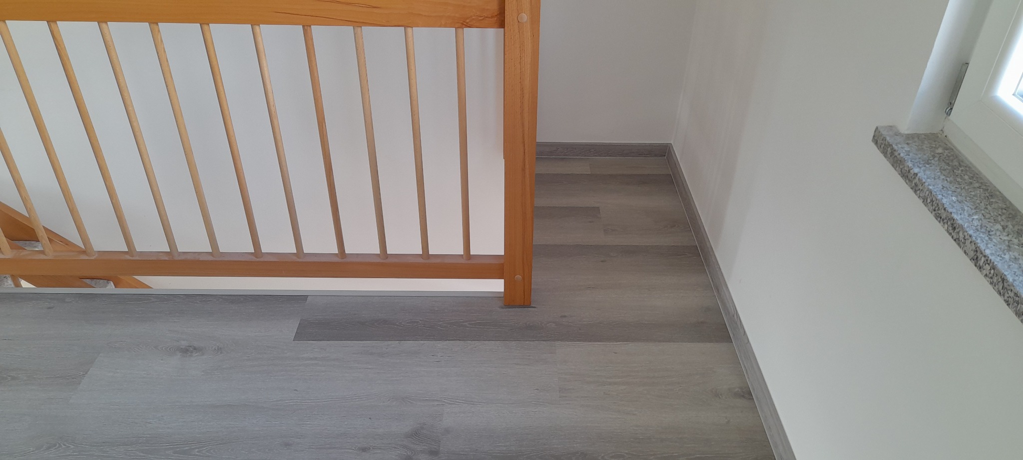 Jasnoszare panele podłogowe ułożone wzdłuż ściany z listwą przypodłogową, fragment drewnianej balustrady schodów i parapet okienny z szarego kamienia.