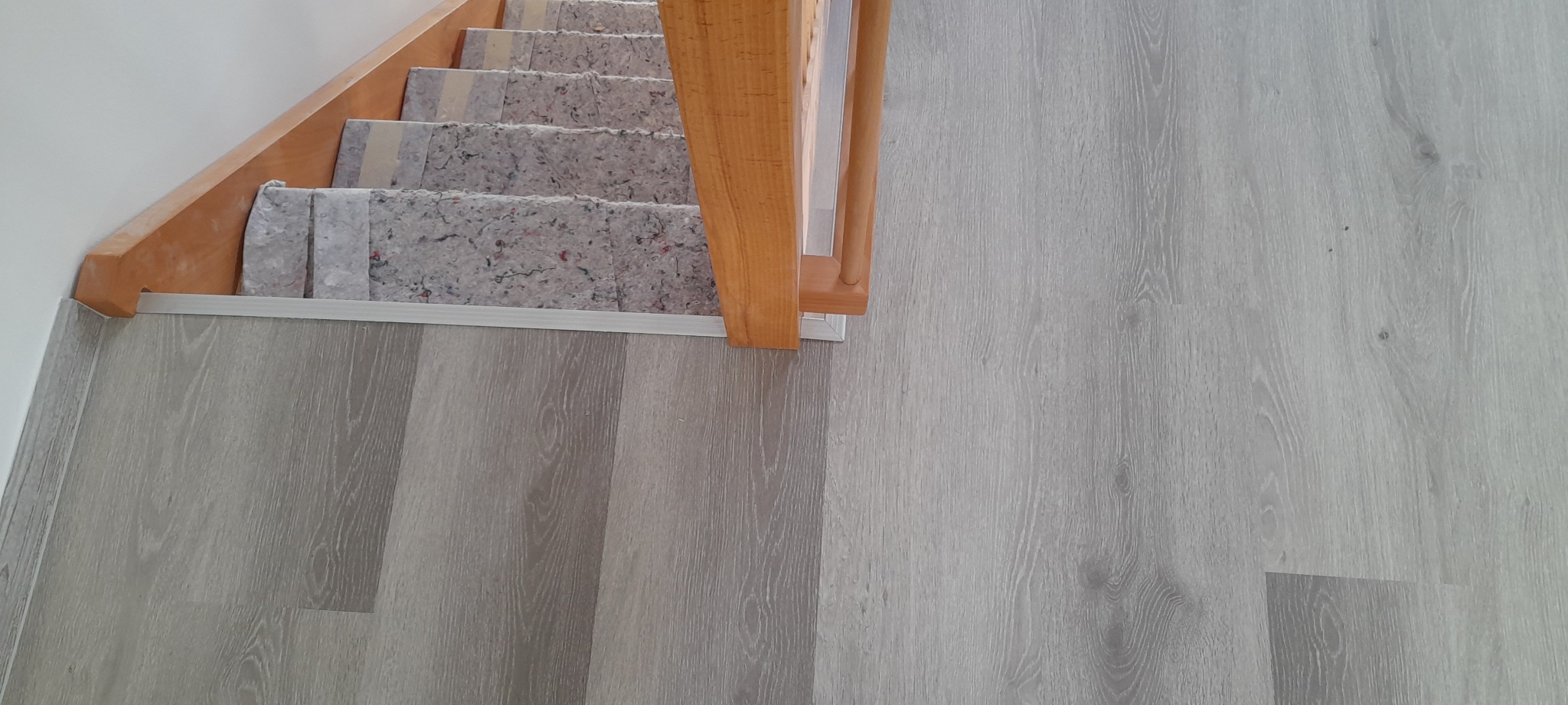 Ułożone panele podłogowe w odcieniach szarości, widoczne schody z drewnianą barierką i podkładem pod dywan, srebrna listwa przypodłogowa.