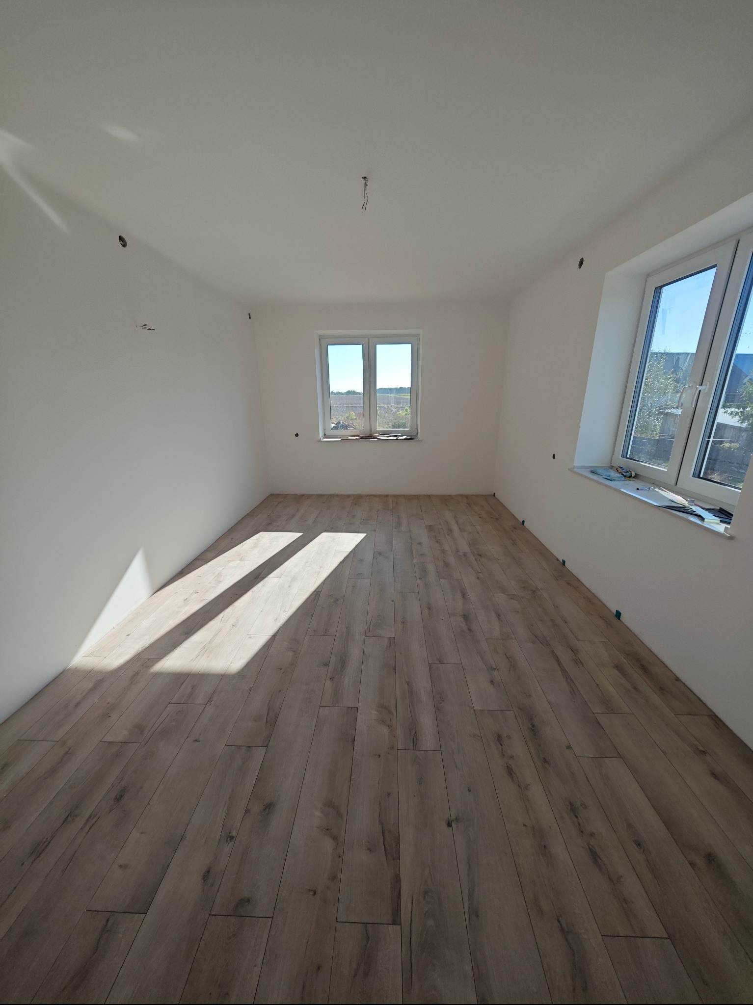 Jasne panele podłogowe w pustym, świeżo wykończonym pokoju z dwoma oknami. Ściany pomalowane na biało, widoczne ślady po instalacji elektrycznej.