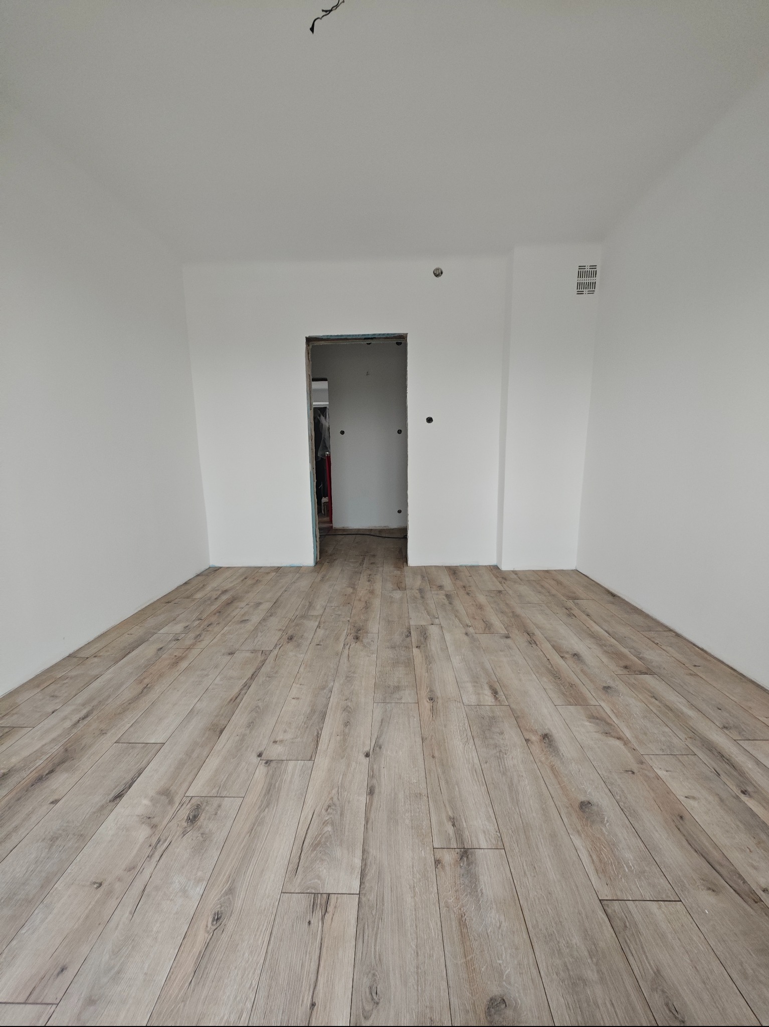 Jasne panele podłogowe ułożone w pustym, świeżo pomalowanym na biało pokoju z widocznym wejściem do kolejnego pomieszczenia. Minimalistyczne wykończenie.