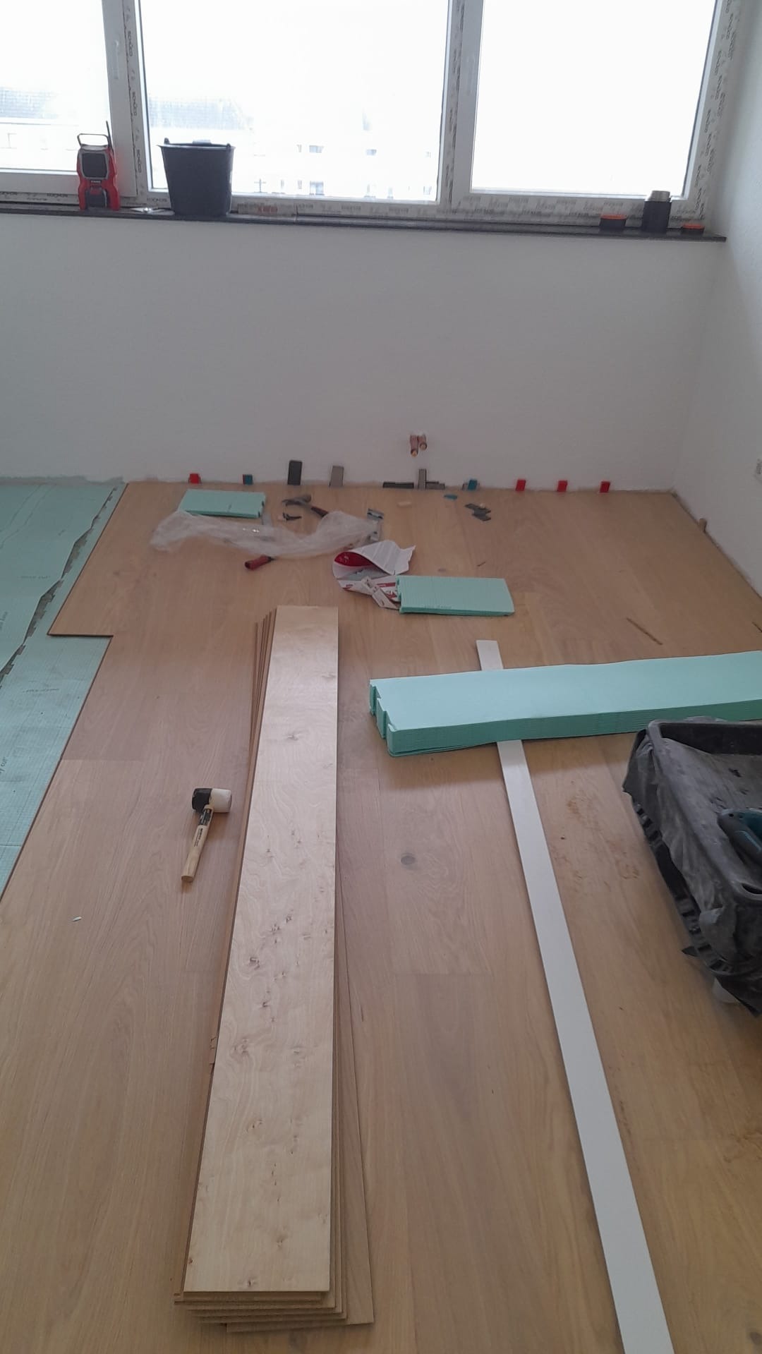 Układanie paneli podłogowych w jasnym wnętrzu. Widoczne panele, podkład, listwy przypodłogowe i narzędzia. Częściowo ułożona podłoga z widocznymi elementami montażowymi.