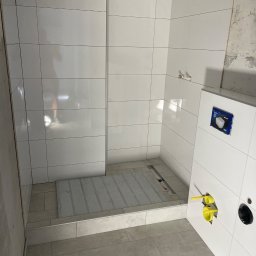 SODELUX - Białe płytki ścienne i podłogowe w trakcie montażu w kabinie prysznicowej, widoczne żółte rury instalacji wodnej i odpływ liniowy w podłodze.