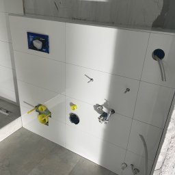 SODELUX - Świeżo położone, białe płytki ścienne w łazience z widocznymi instalacjami hydraulicznymi i elektrycznymi w trakcie montażu, szare płytki podłogowe, surowy beton na górze ściany.