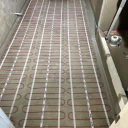 SODELUX - Instalacja ogrzewania podłogowego w łazience, widoczne czerwone kable grzewcze na siatce montażowej, przygotowane do zalania wylewką samopoziomującą.