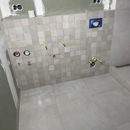 SODELUX - Świeżo wykafelkowana ściana w łazience z przygotowanymi otworami na instalacje wodno-kanalizacyjne i elektryczne; widoczne puszki pod gniazdka, rury doprowadzające wodę, oraz fragment ściany...