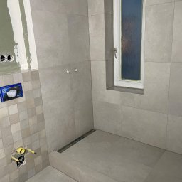 SODELUX - Szara łazienka w trakcie remontu: widoczne płytki ścienne i podłogowe, okno wnękowe, przygotowane instalacje wodno-kanalizacyjne, oraz częściowo wykończona ściana z elementami gipsowania.