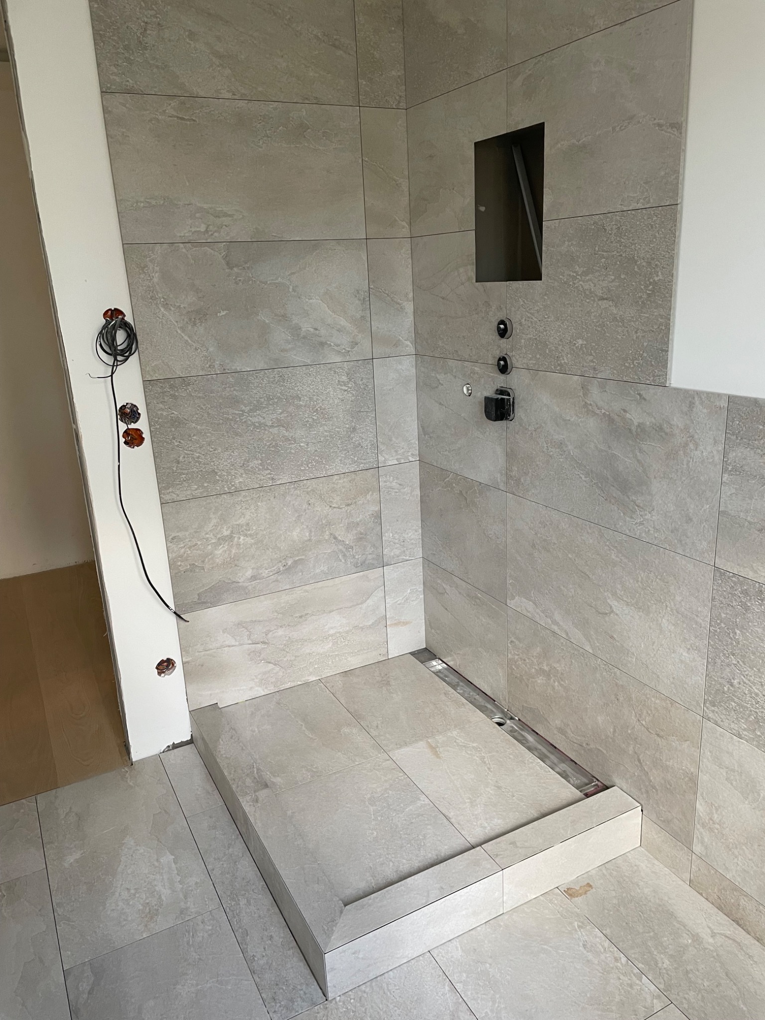 Niedokończona kabina prysznicowa z odpływem liniowym i wnęką w ścianie, widoczne przewody elektryczne i niedoróbki przy wykończeniu.