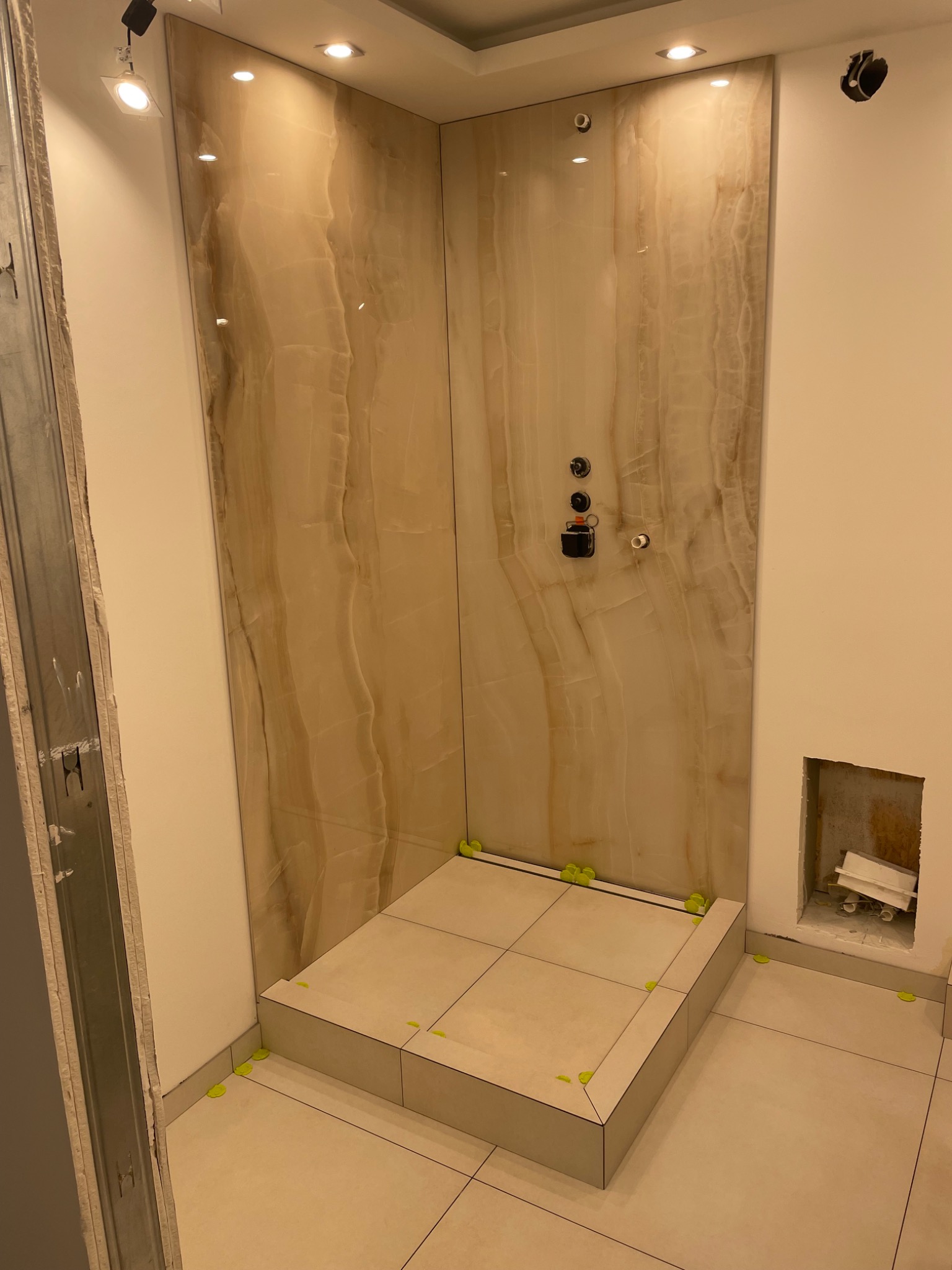 Fragment łazienki w trakcie prac wykończeniowych: ułożone płytki na podłodze i ścianie prysznica z imitacją marmuru, widoczne dystanse krzyżykowe, przygotowane otwory na armaturę oraz niedokończona...