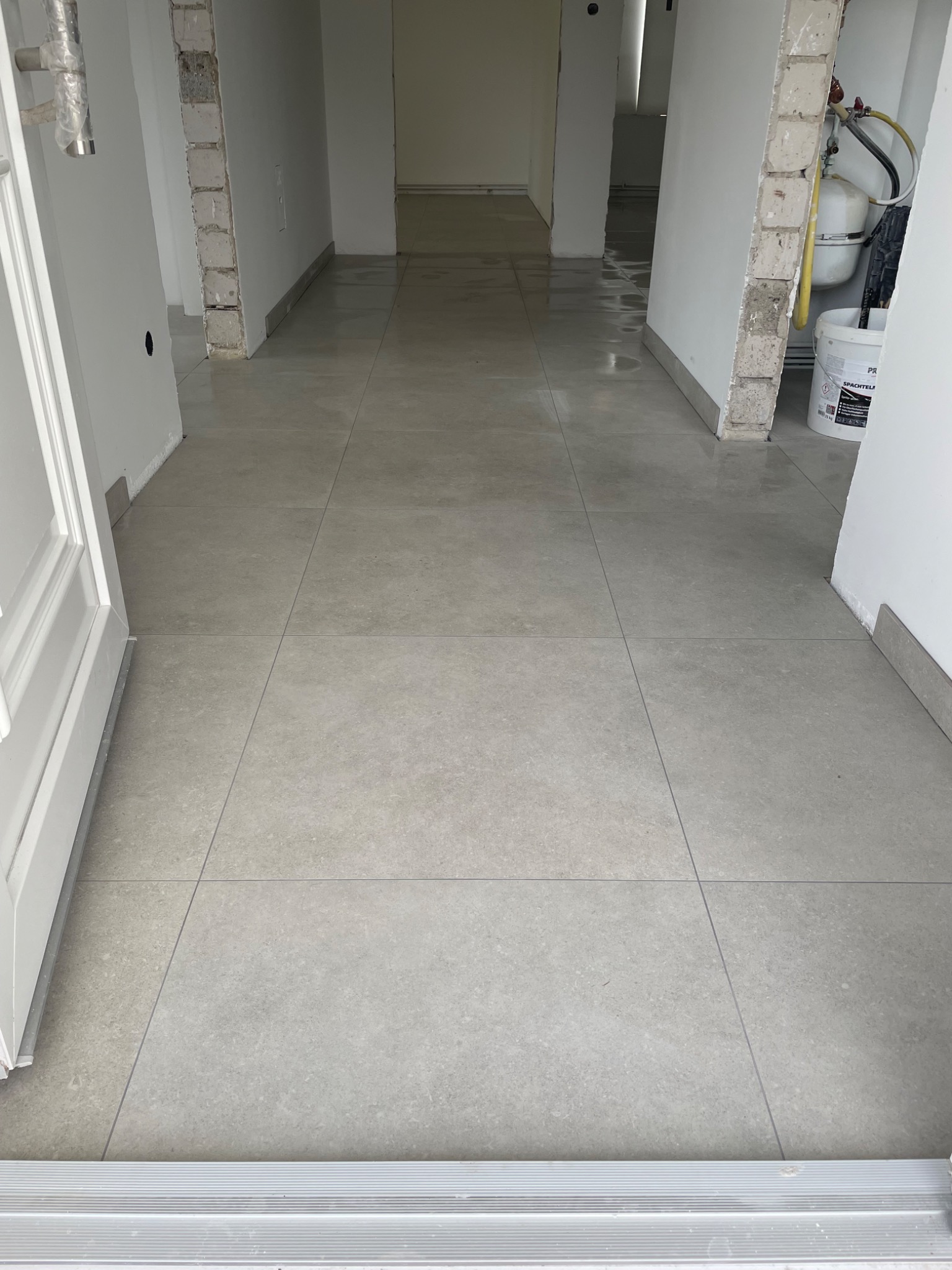Szare, kwadratowe płytki podłogowe ułożone w korytarzu podczas remontu, widoczny otwarty pokój w tle i wiadro z zaprawą na ścianie po prawej stronie.