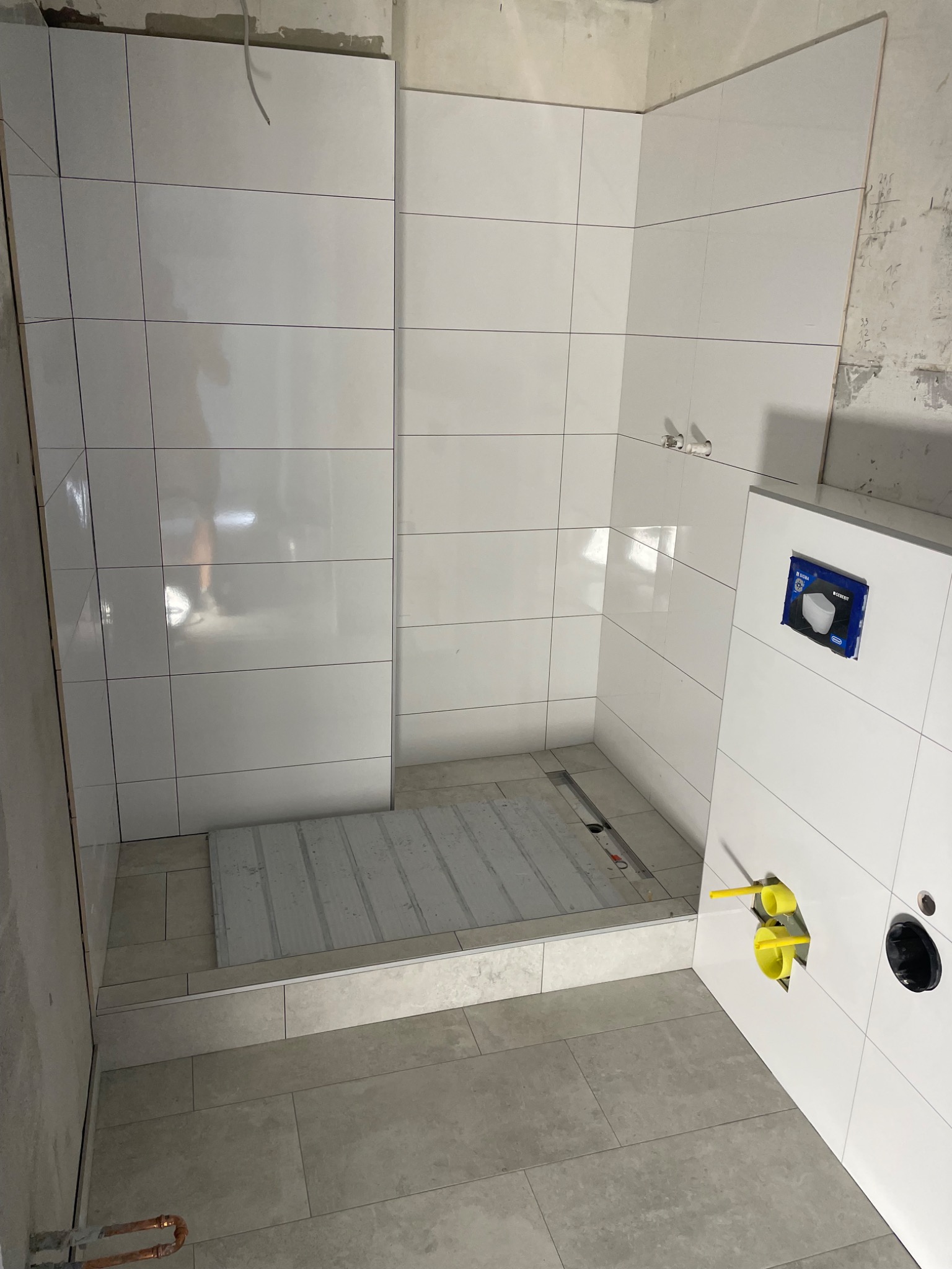 Białe płytki ścienne i podłogowe w trakcie montażu w kabinie prysznicowej, widoczne żółte rury instalacji wodnej i odpływ liniowy w podłodze.