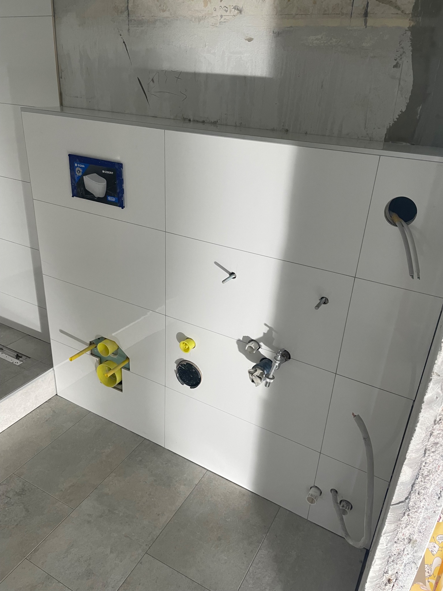 Świeżo położone, białe płytki ścienne w łazience z widocznymi instalacjami hydraulicznymi i elektrycznymi w trakcie montażu, szare płytki podłogowe, surowy beton na górze ściany.