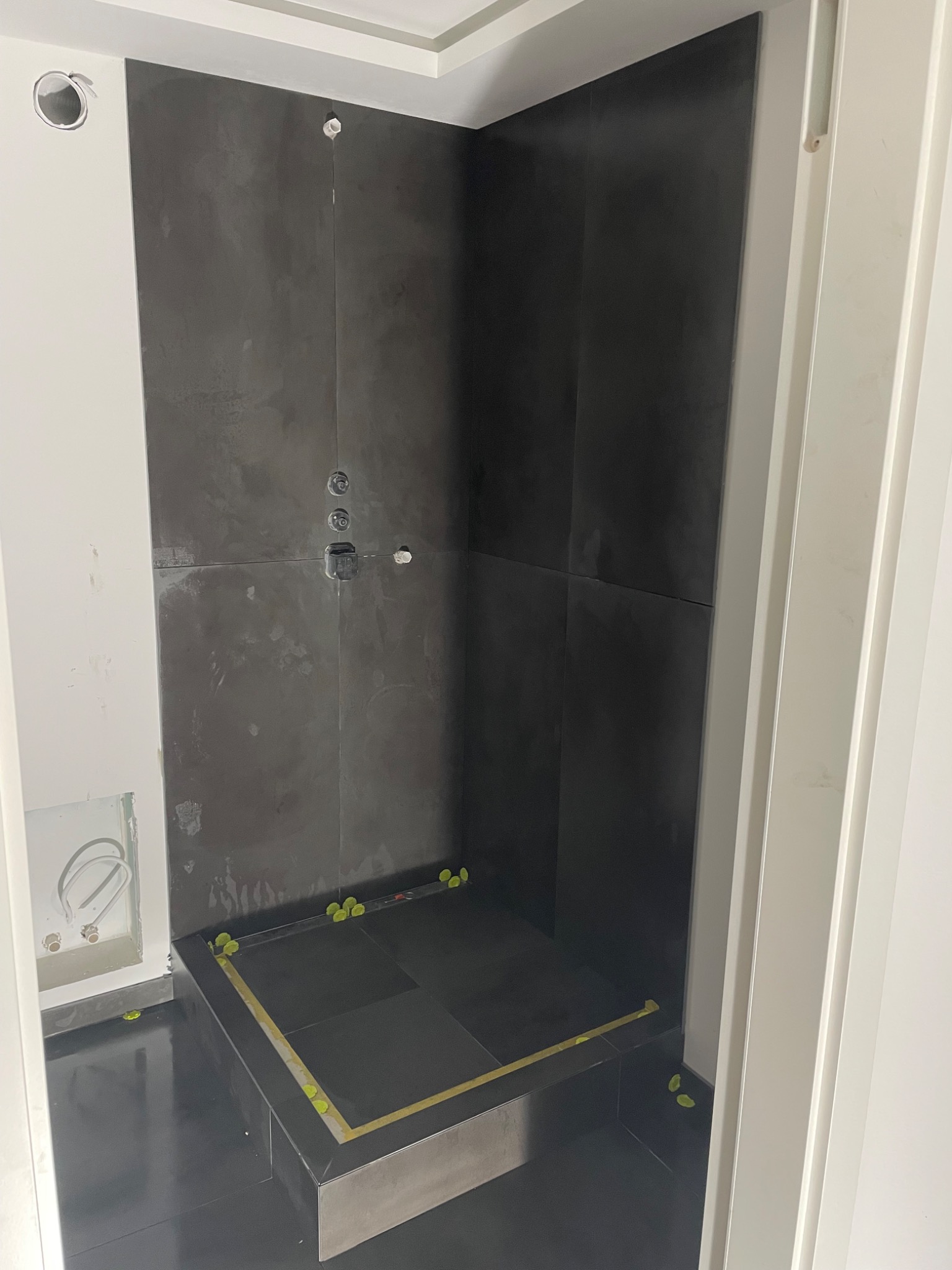 Montaż ciemnych, matowych płytek w kabinie prysznicowej, widoczne krzyżyki glazurnicze i wyprowadzone instalacje wodne.