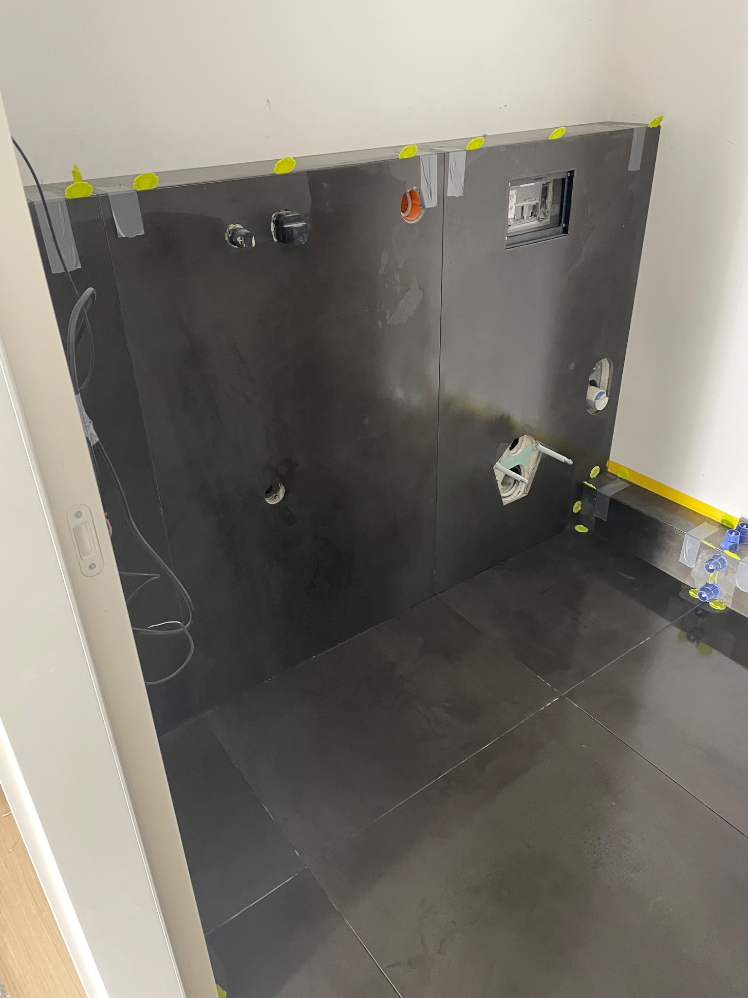Ciemne, błyszczące płytki na ścianie i podłodze w trakcie montażu, widoczne otwory na armaturę, szara taśma i żółte kliny poziomujące płytki.