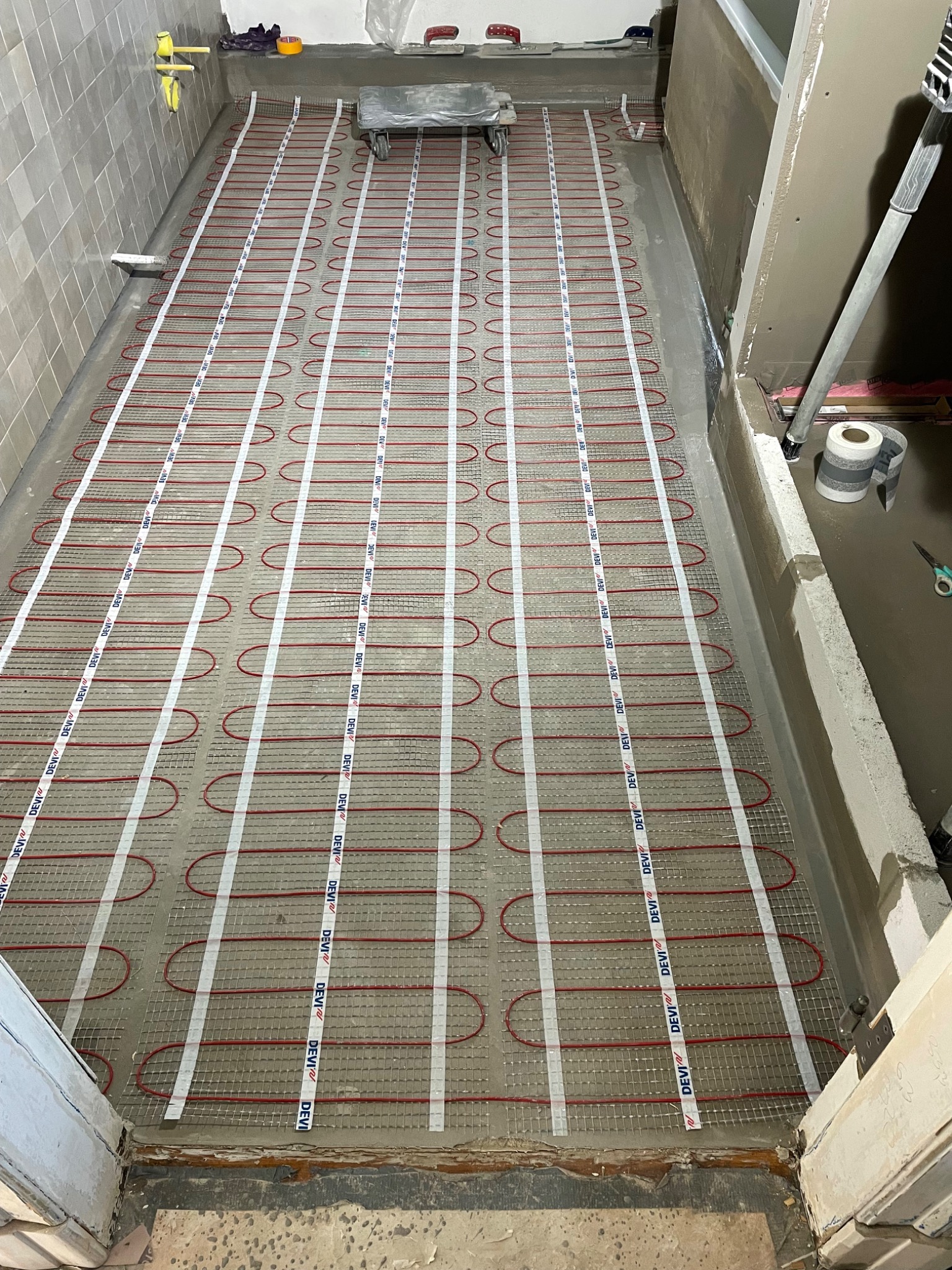 Instalacja ogrzewania podłogowego w łazience, widoczne czerwone kable grzewcze na siatce montażowej, przygotowane do zalania wylewką samopoziomującą.