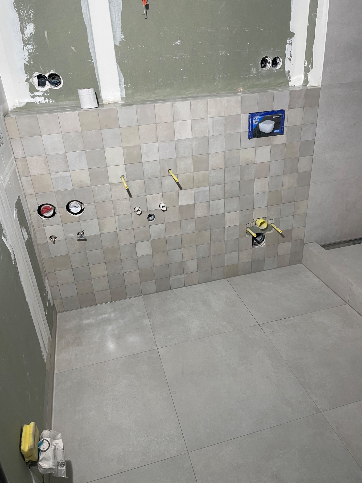 Świeżo wykafelkowana ściana w łazience z przygotowanymi otworami na instalacje wodno-kanalizacyjne i elektryczne; widoczne puszki pod gniazdka, rury doprowadzające wodę, oraz fragment ściany...