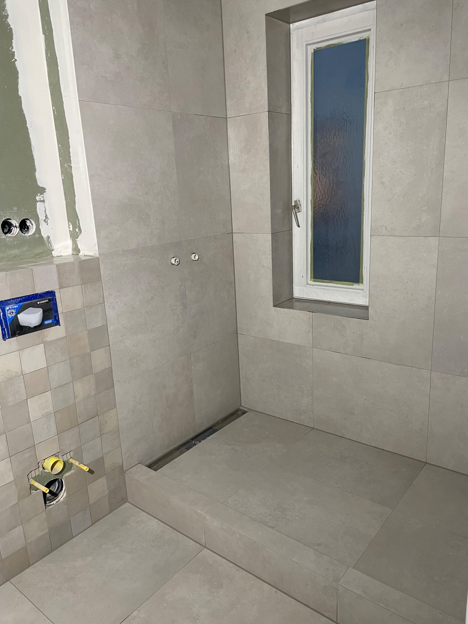 Szara łazienka w trakcie remontu: widoczne płytki ścienne i podłogowe, okno wnękowe, przygotowane instalacje wodno-kanalizacyjne, oraz częściowo wykończona ściana z elementami gipsowania.