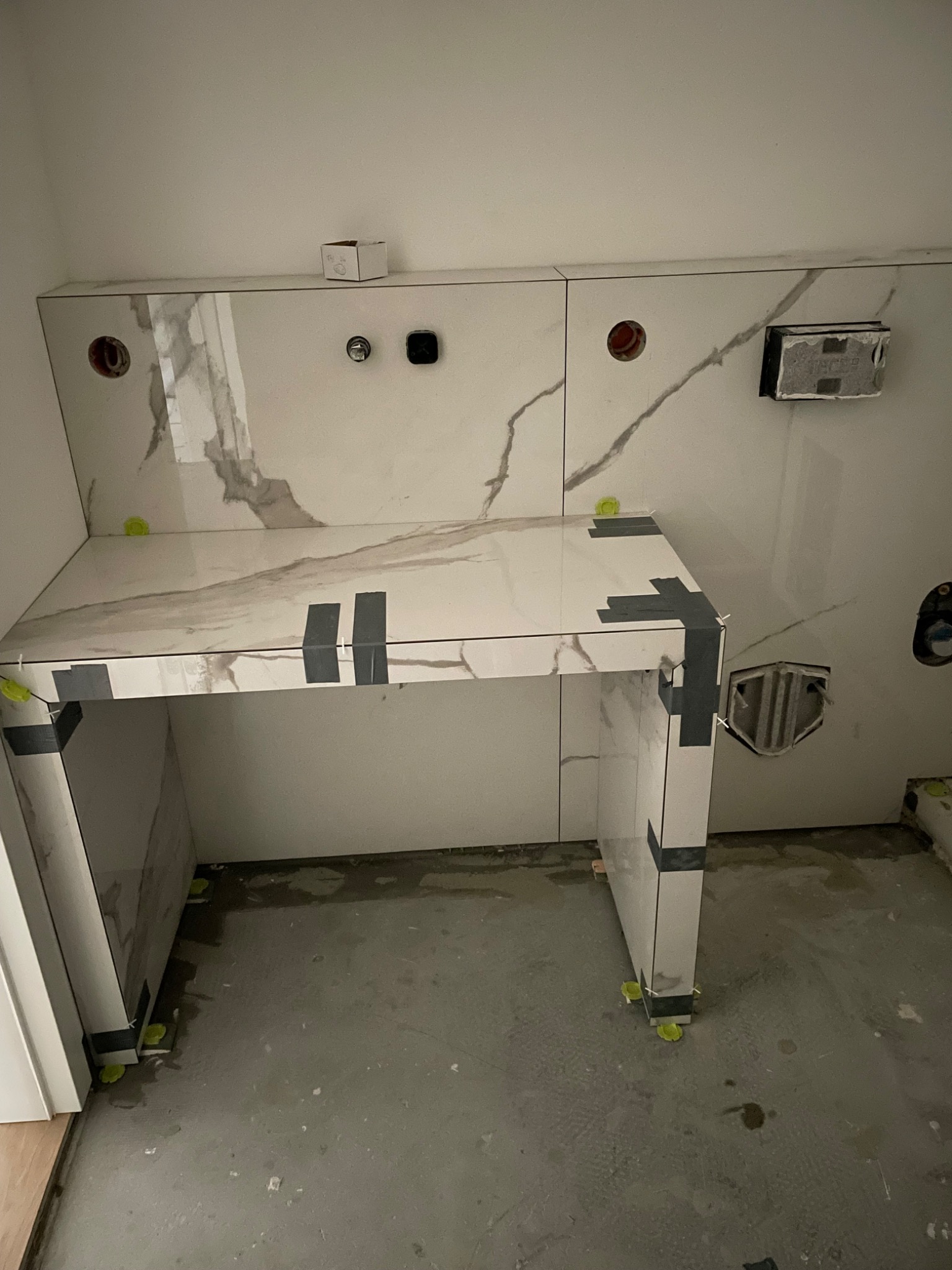 W trakcie prac: zabudowa z płytek imitujących marmur w łazience w Zabrzu, zabezpieczona taśmą malarską i poziomowana klinami, widoczne otwory na instalacje.