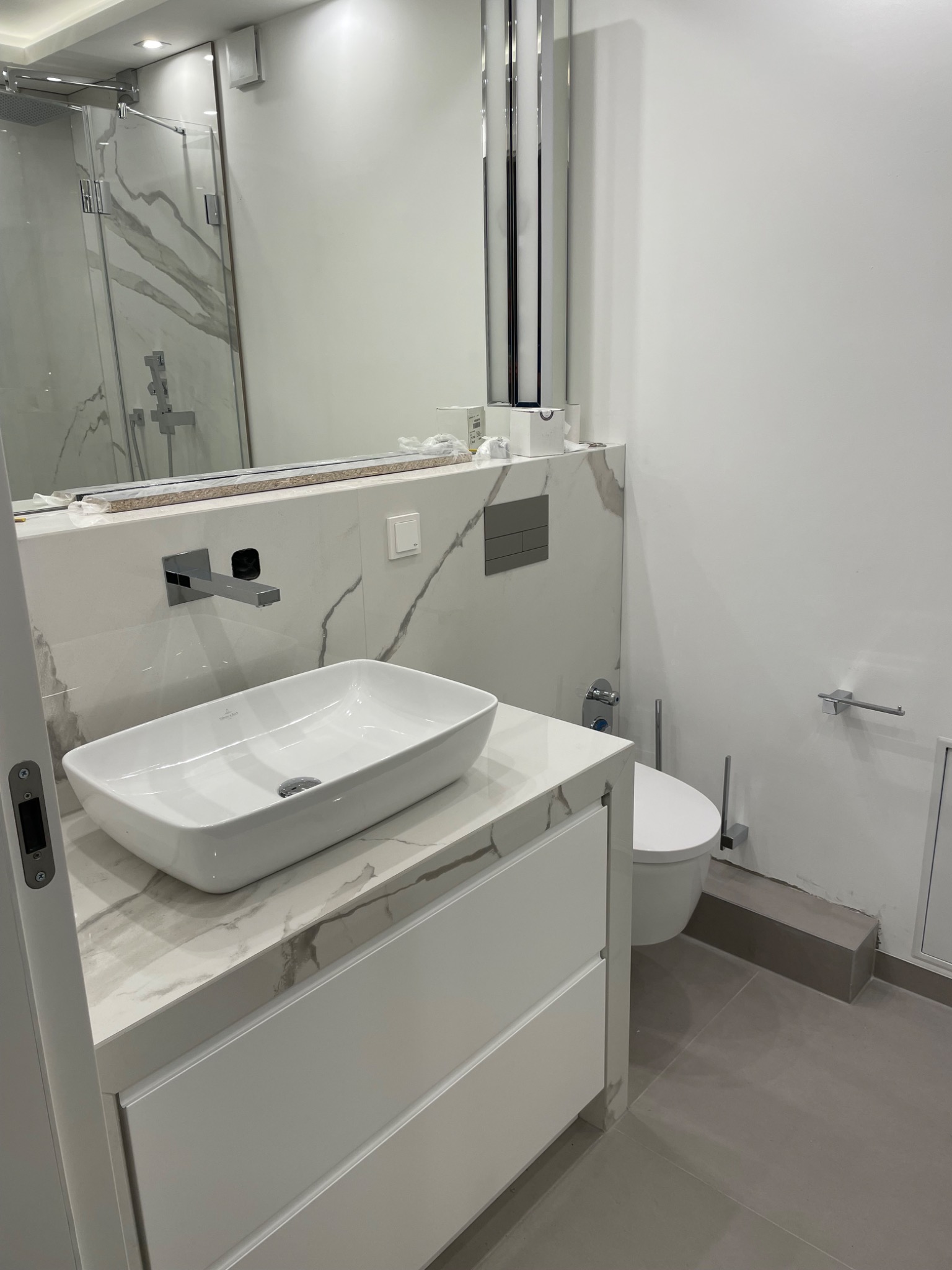 Nowoczesna łazienka w trakcie wykańczania: biała umywalka nablatowa Villeroy & Boch na szafce z blatem imitującym marmur, wisząca toaleta z szarą płytką przycisku, szklana kabina prysznicowa w tle,...