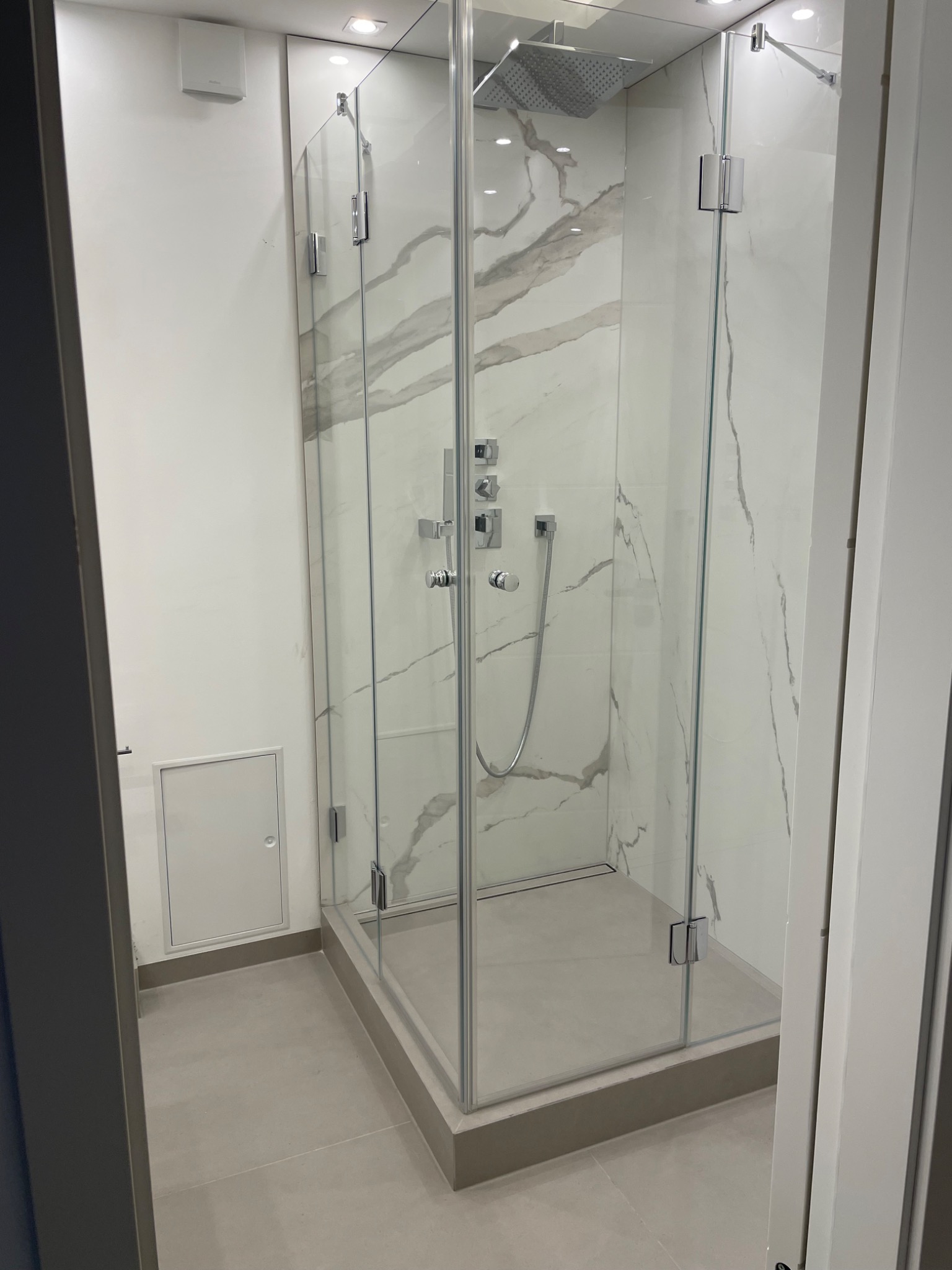 Nowoczesna kabina prysznicowa z deszczownicą i marmurowymi płytkami, szklane drzwi z chromowanymi zawiasami, minimalistyczny design, widoczna kratka ściekowa.