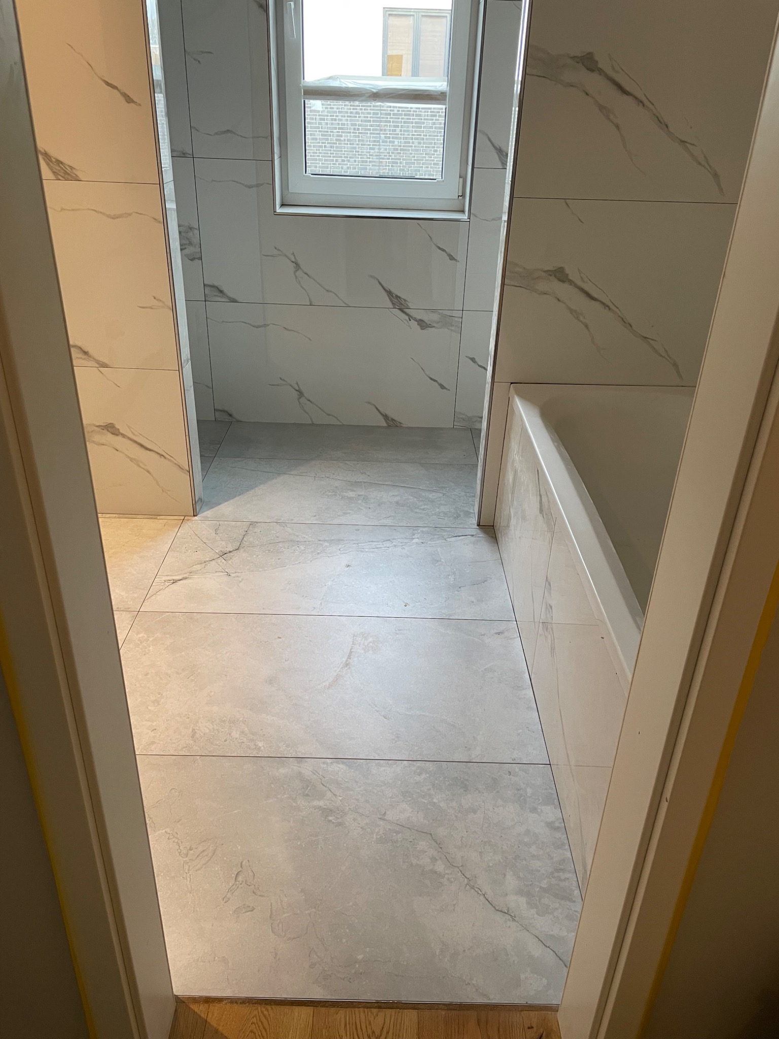 Wykończona łazienka z płytkami imitującymi marmur na ścianach i podłodze, widok zza progu, biała wanna po prawej stronie i okno w tle.