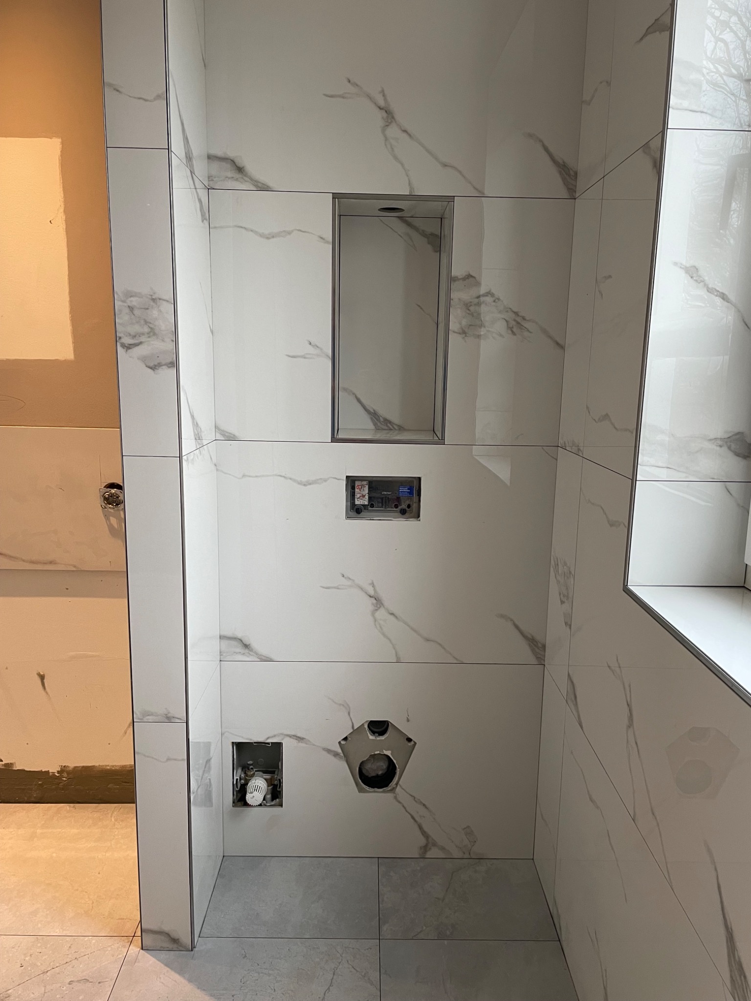 Ściana łazienki w trakcie wykańczania, z białymi płytkami imitującymi marmur, wnęką na kosmetyki, widocznymi instalacjami hydraulicznymi i częściowo wykończoną ścianą obok.