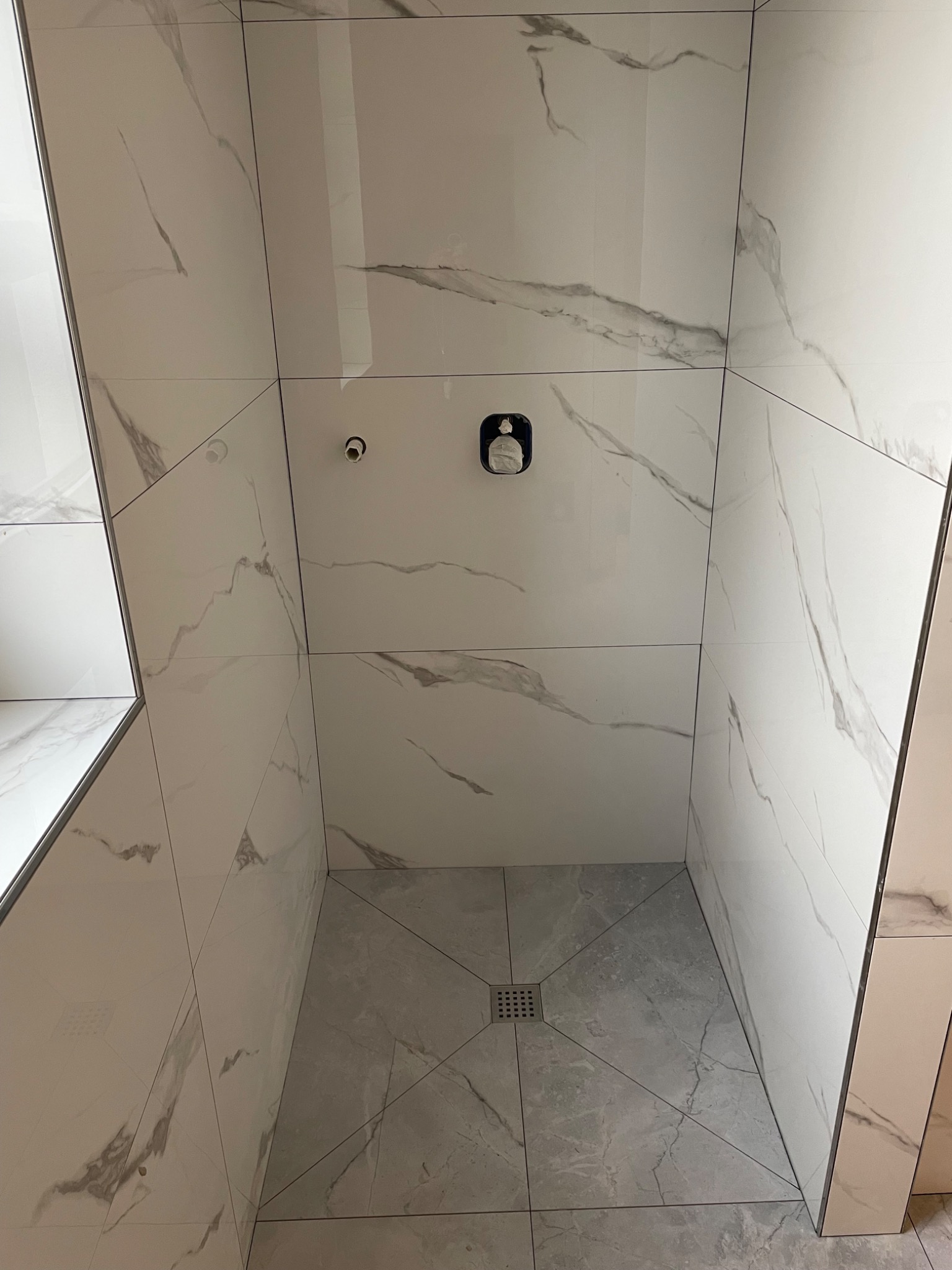 Wnętrze kabiny prysznicowej wyłożonej białymi płytkami z szarym marmurkowym wzorem, widoczny odpływ liniowy w podłodze i zamontowana bateria prysznicowa.