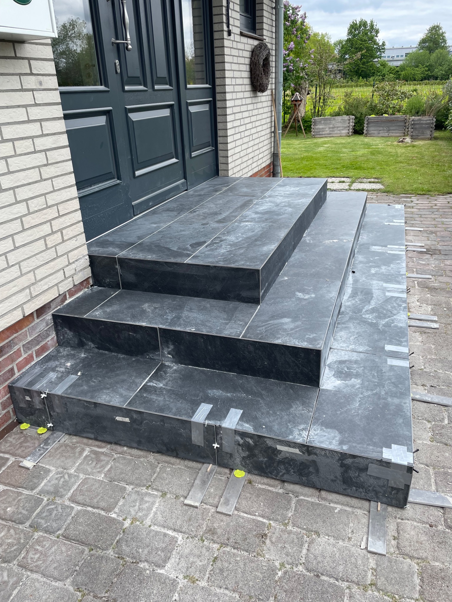 Wyłożone ciemnoszarą płytką schody wejściowe do domu, zabezpieczone taśmą i listwami dystansowymi, widoczne elementy elewacji z cegły i kostki brukowej.